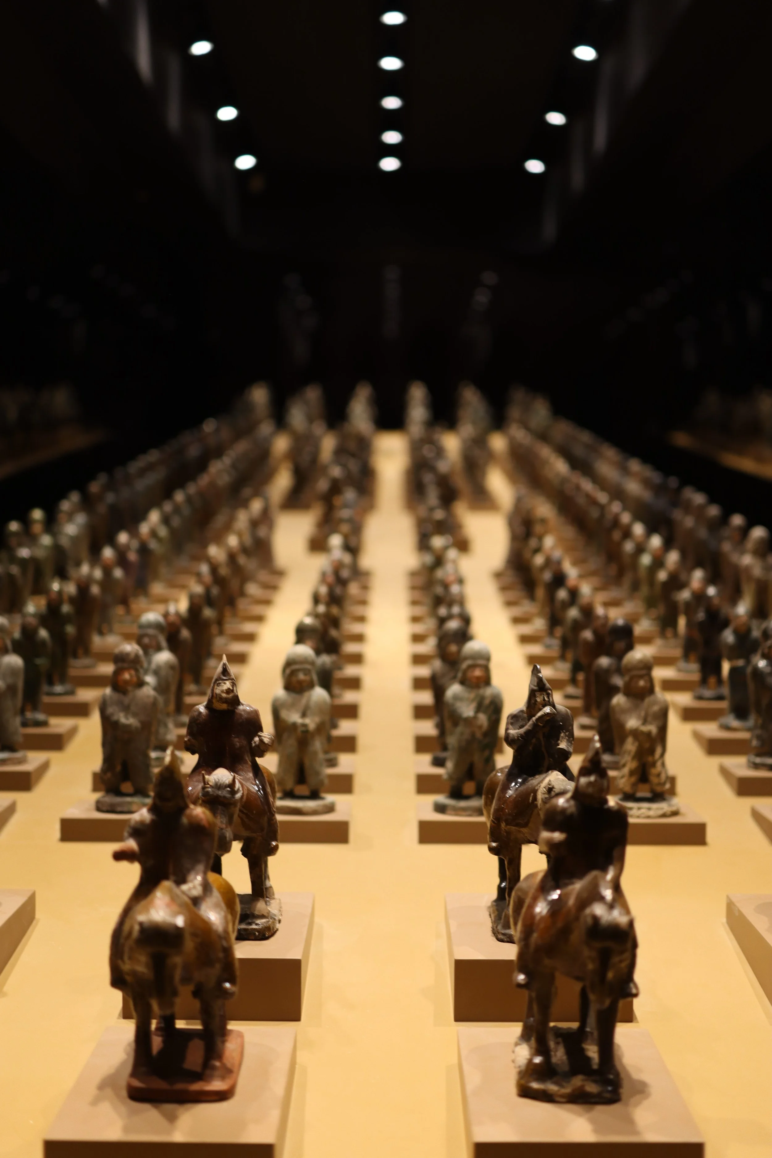 Display of numerous small, ancient clay figurines of warriors, arranged in rows on a yellow surface inside a dimly lit museum exhibit.