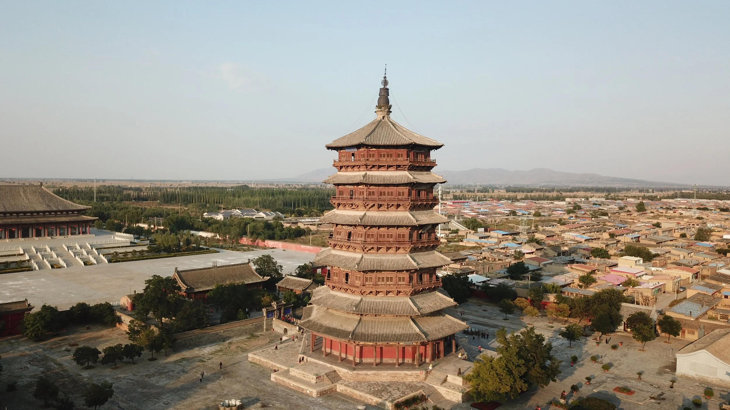 An ancient multi-tiered pagoda with traditional Chinese architecture, surrounded by smaller buildings and trees, in a historic site with a city and distant mountains in the background.