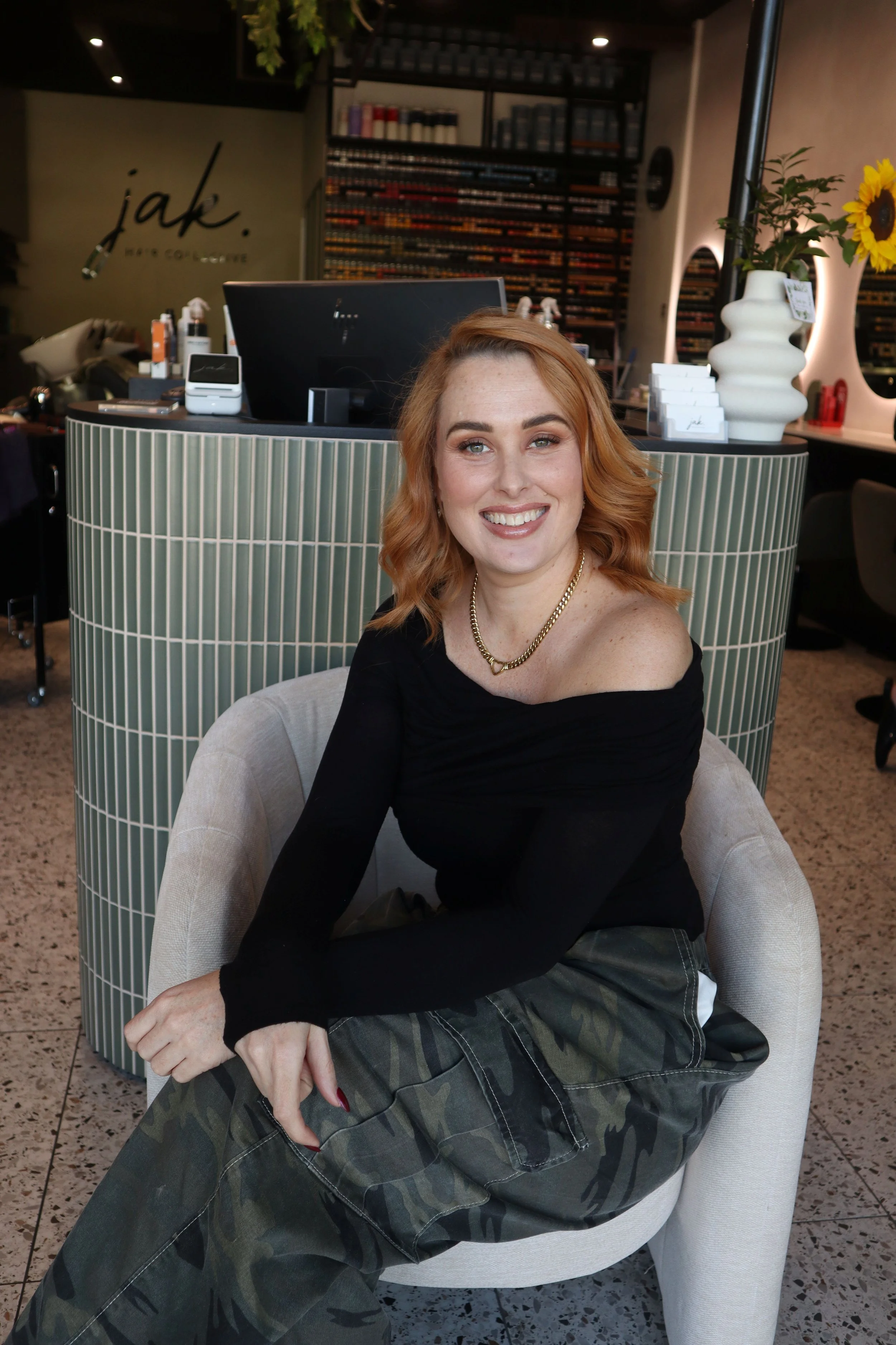 A woman with shoulder-length red hair, wearing a black dress with semi-transparent, polka-dotted sleeves, sitting on a green chair with a gold frame in what appears to be a cafe or restaurant. She is smiling at the camera, with her hands clasped in her lap, and has a watch on her left wrist.