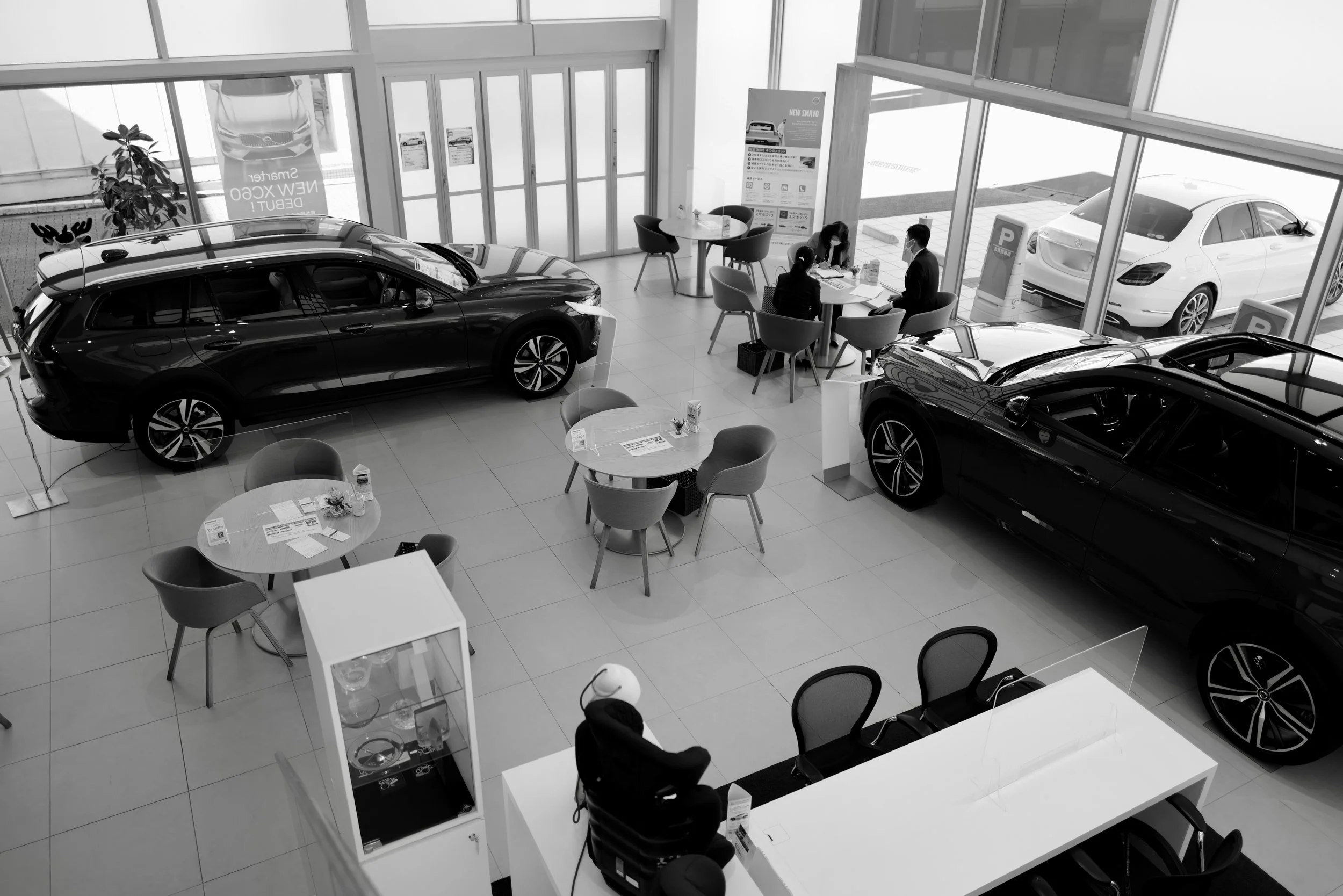 Black and white photo of a car dealership showroom with several cars on display, customer seating area with tables and chairs, and a few people, including sales staff, interacting at a table near large windows with parked cars outside.