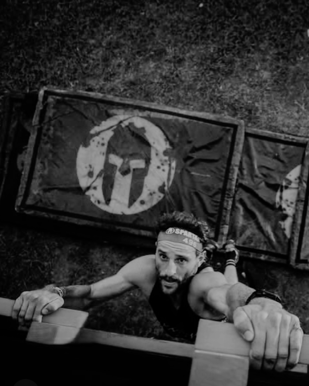 A man wearing a headband and athletic attire looks up at the camera while climbing a wooden wall during an obstacle course, with a flag featuring a Spartan logo on the ground.