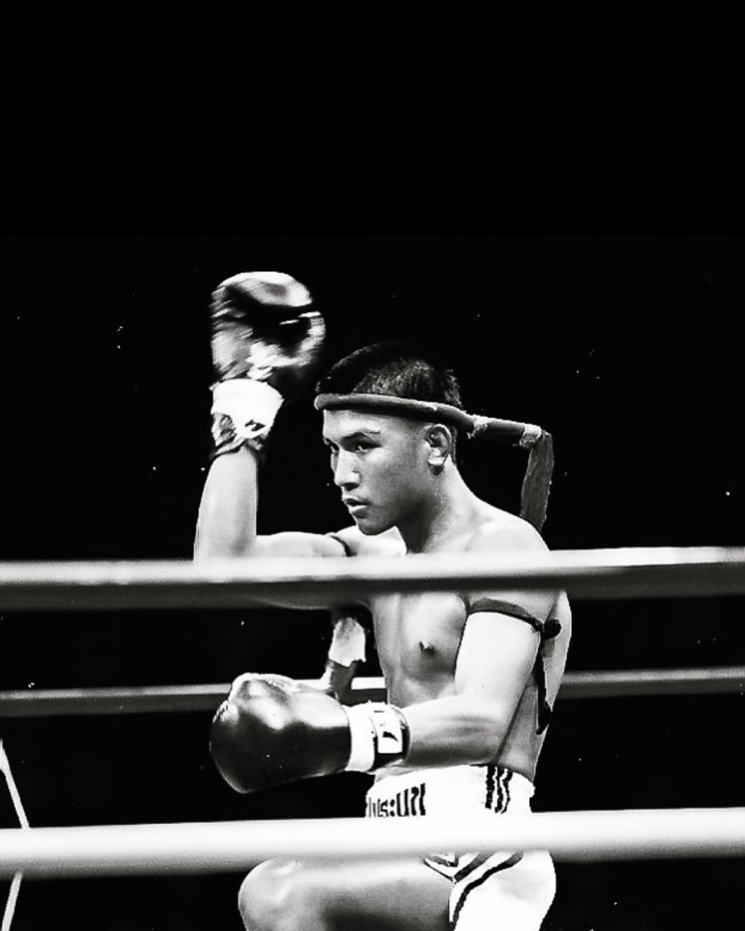 Black-and-white photo of a male boxer in a boxing ring, wearing boxing gloves, shorts, and a headband, with a focused expression.