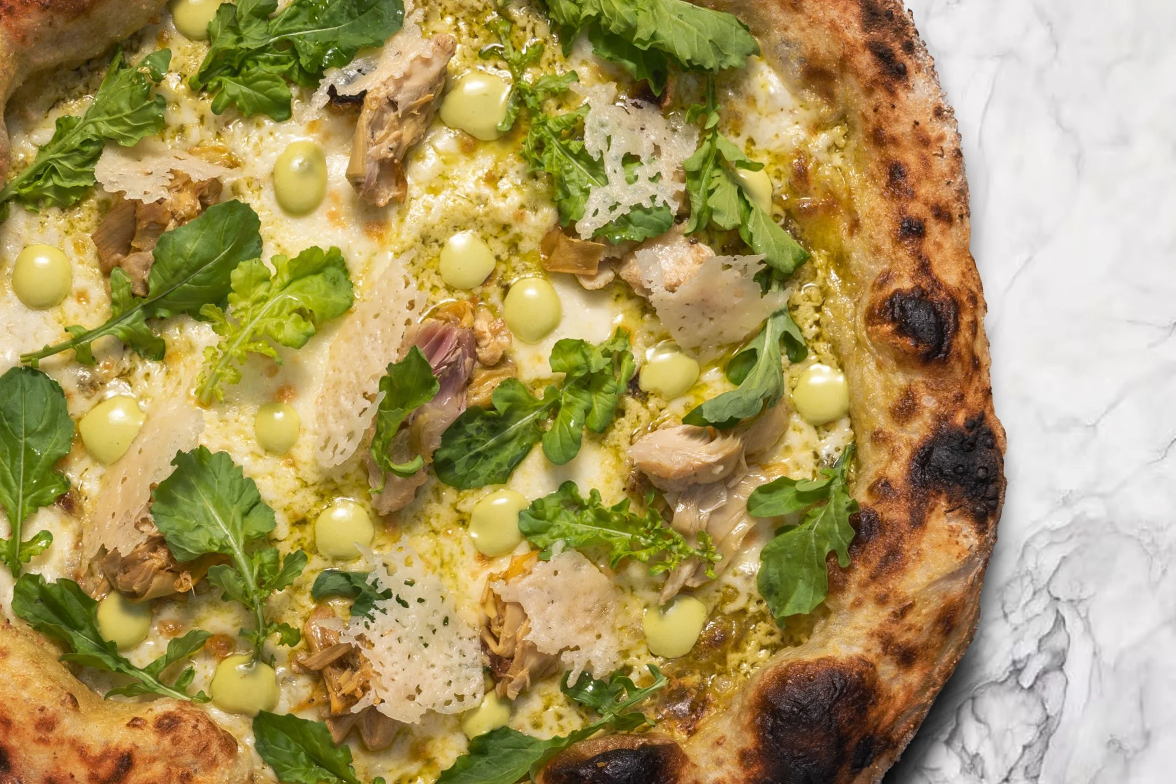 Close-up of a pizza with cheese, greens, chicken, and cheese shavings on a paper surface.