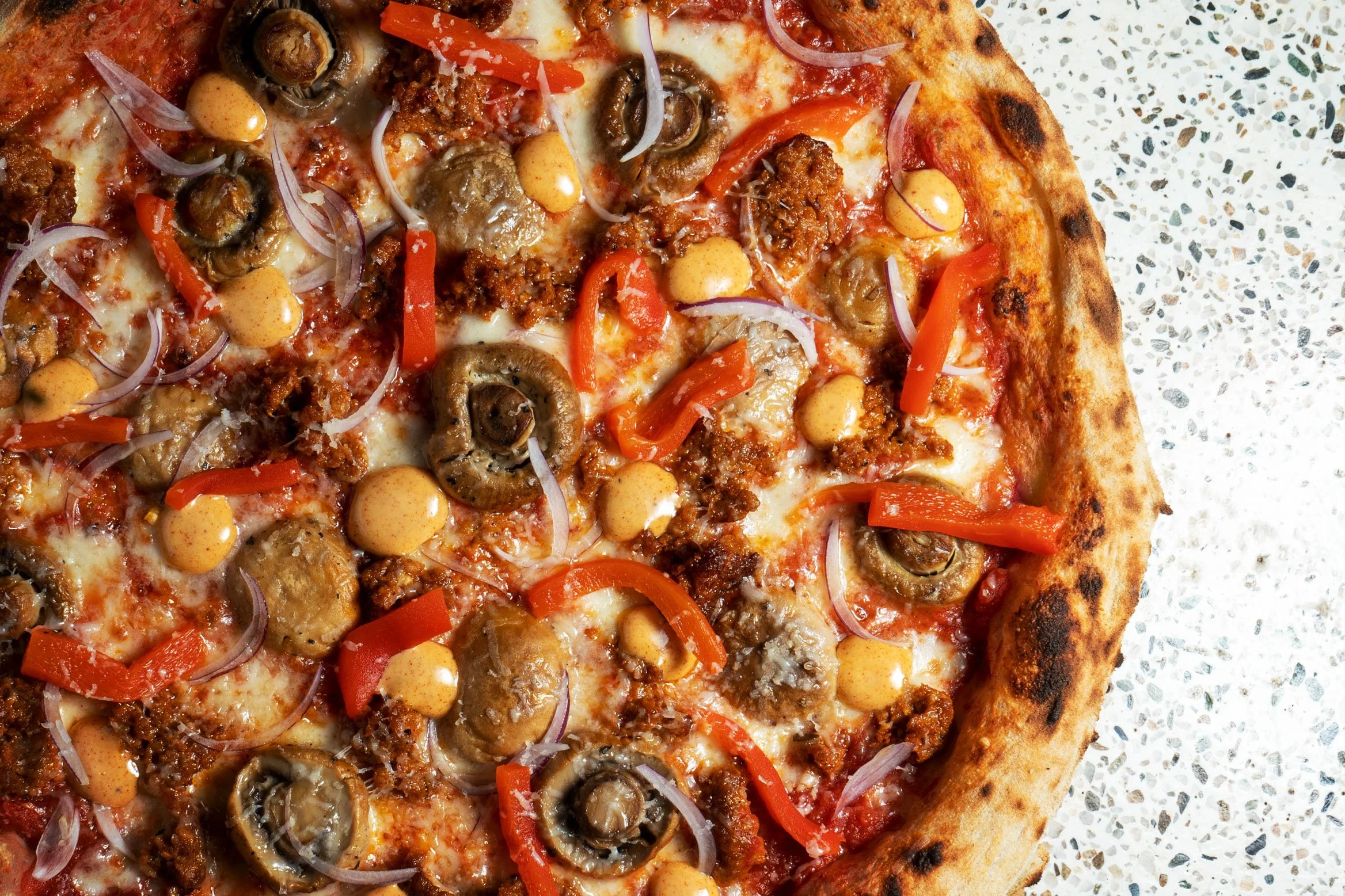 Close-up of a pizza with cheese, mushrooms, red bell peppers, onions, ground beef, and hot sauce on a speckled surface.