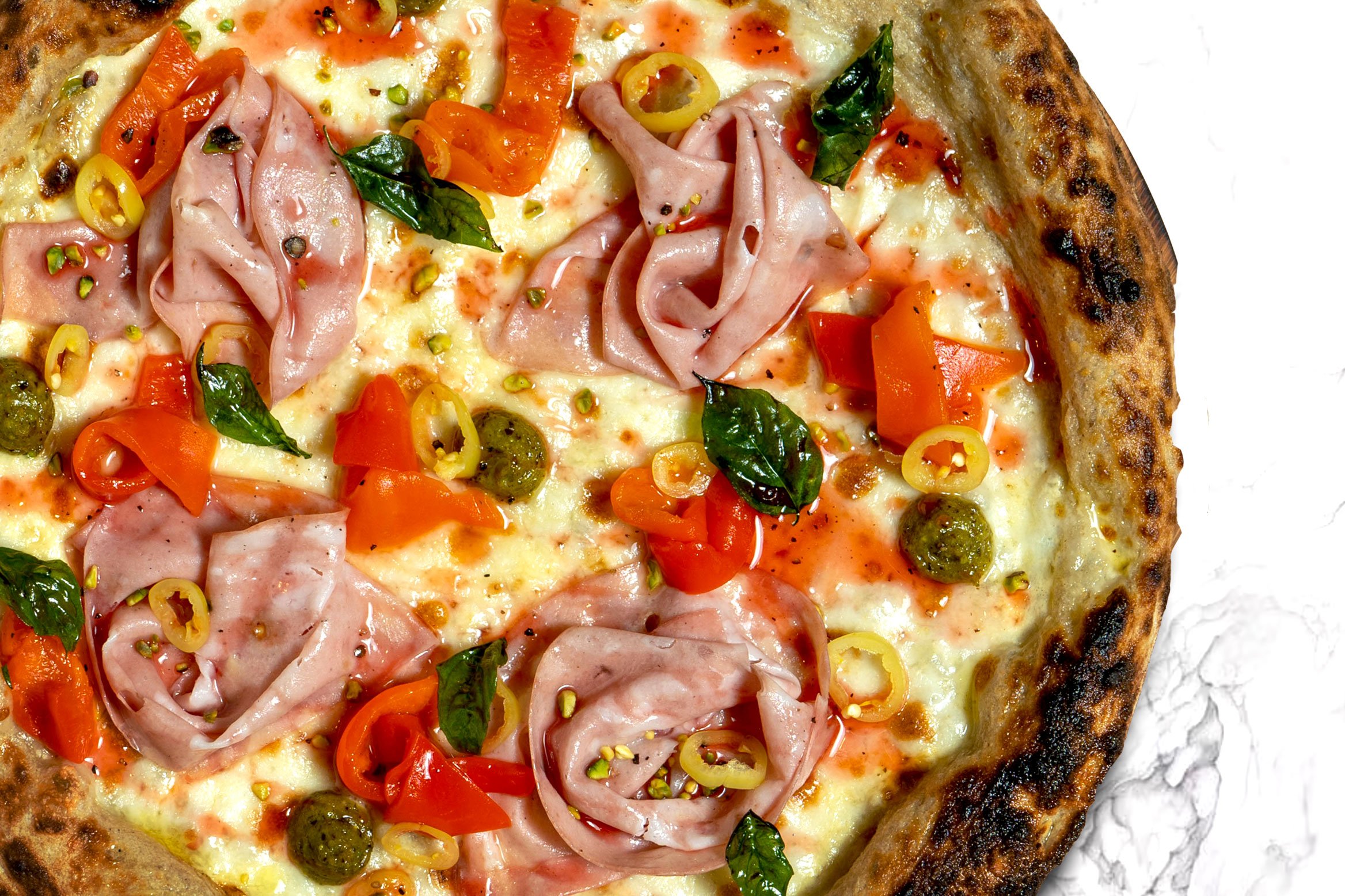 Close-up of a pizza with ham, red and yellow bell peppers, green olives, fresh basil, and sliced yellow chili peppers on a thin crust.