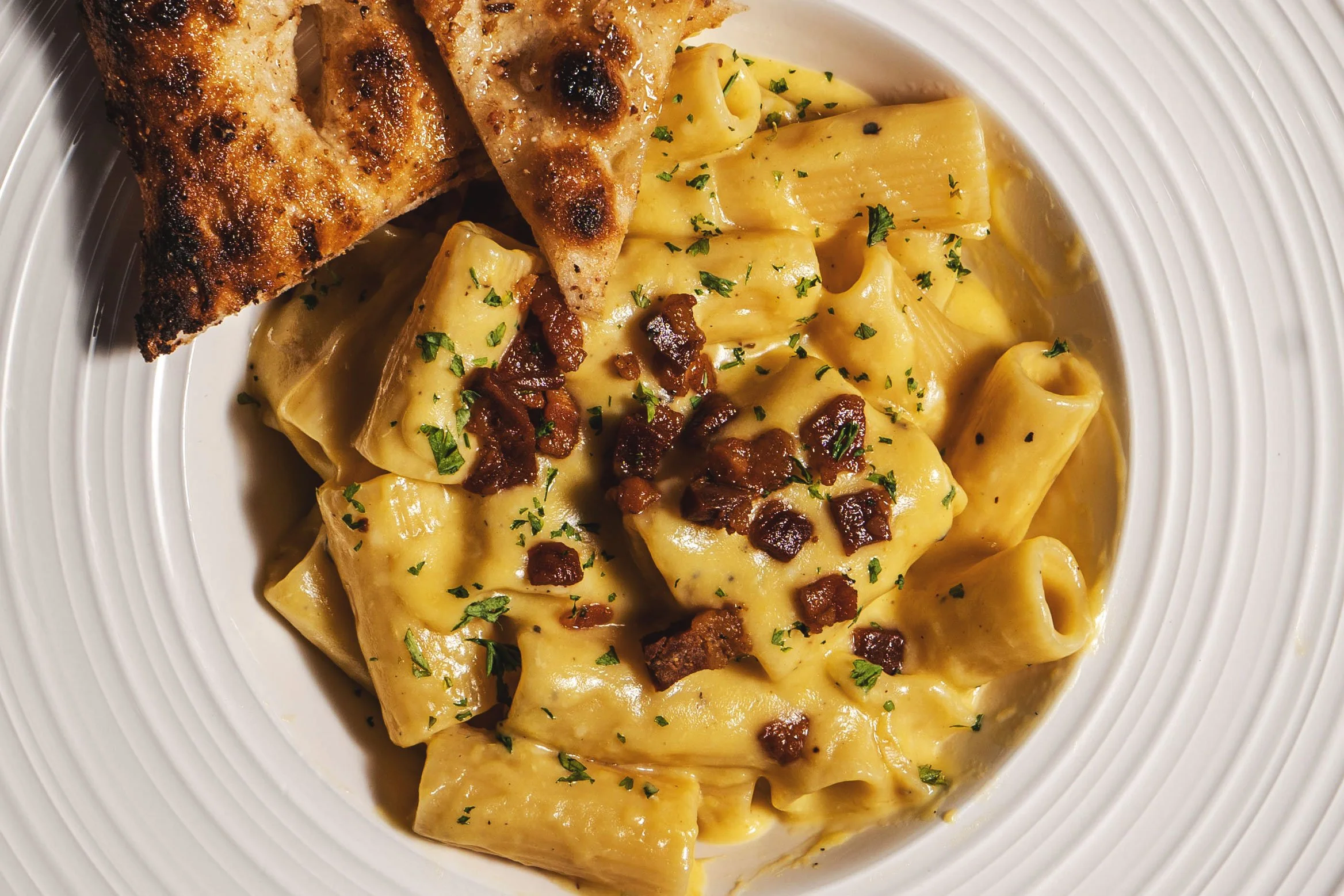A white plate with tortellini pasta in creamy sauce topped with chopped herbs and crispy bacon bits, served with a toasted bread triangle.