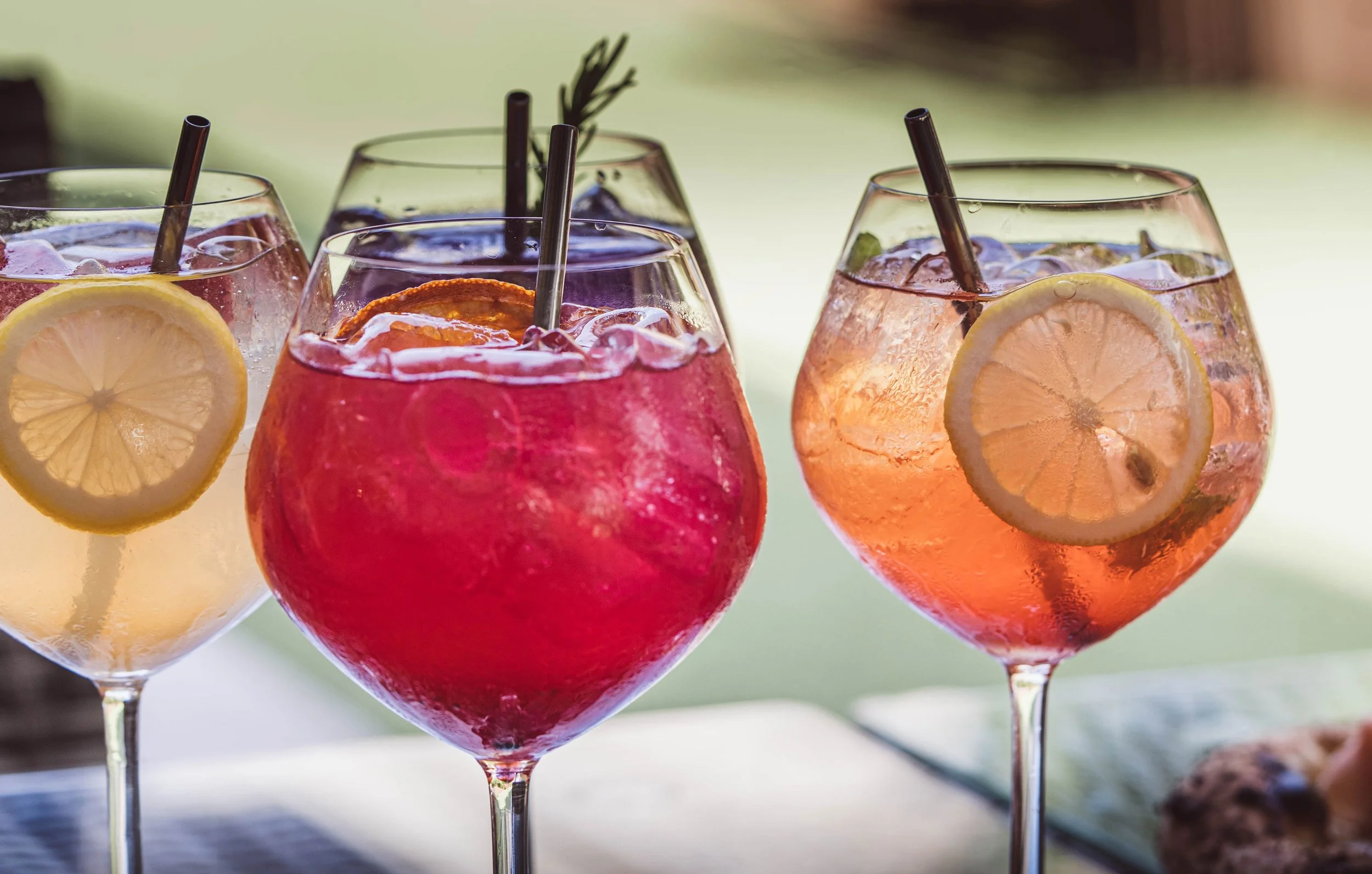 Four colorful cocktails in stemmed glasses with lemon slices and ice, set outdoors.