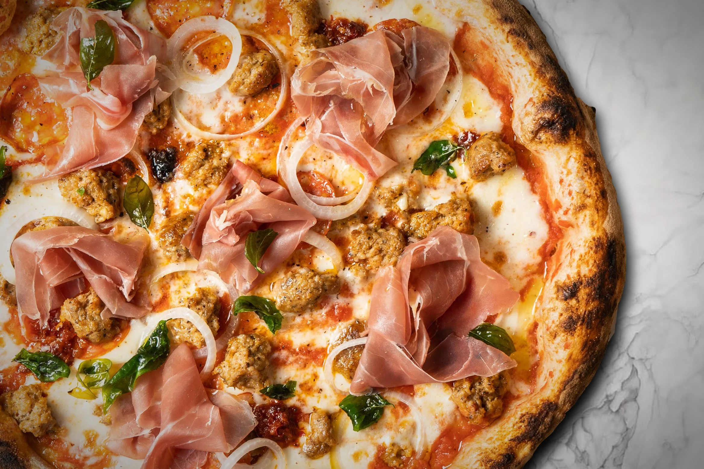 Close-up of a pizza topped with prosciutto, sausage, onion slices, basil, cheese, and tomato sauce on a marble surface.