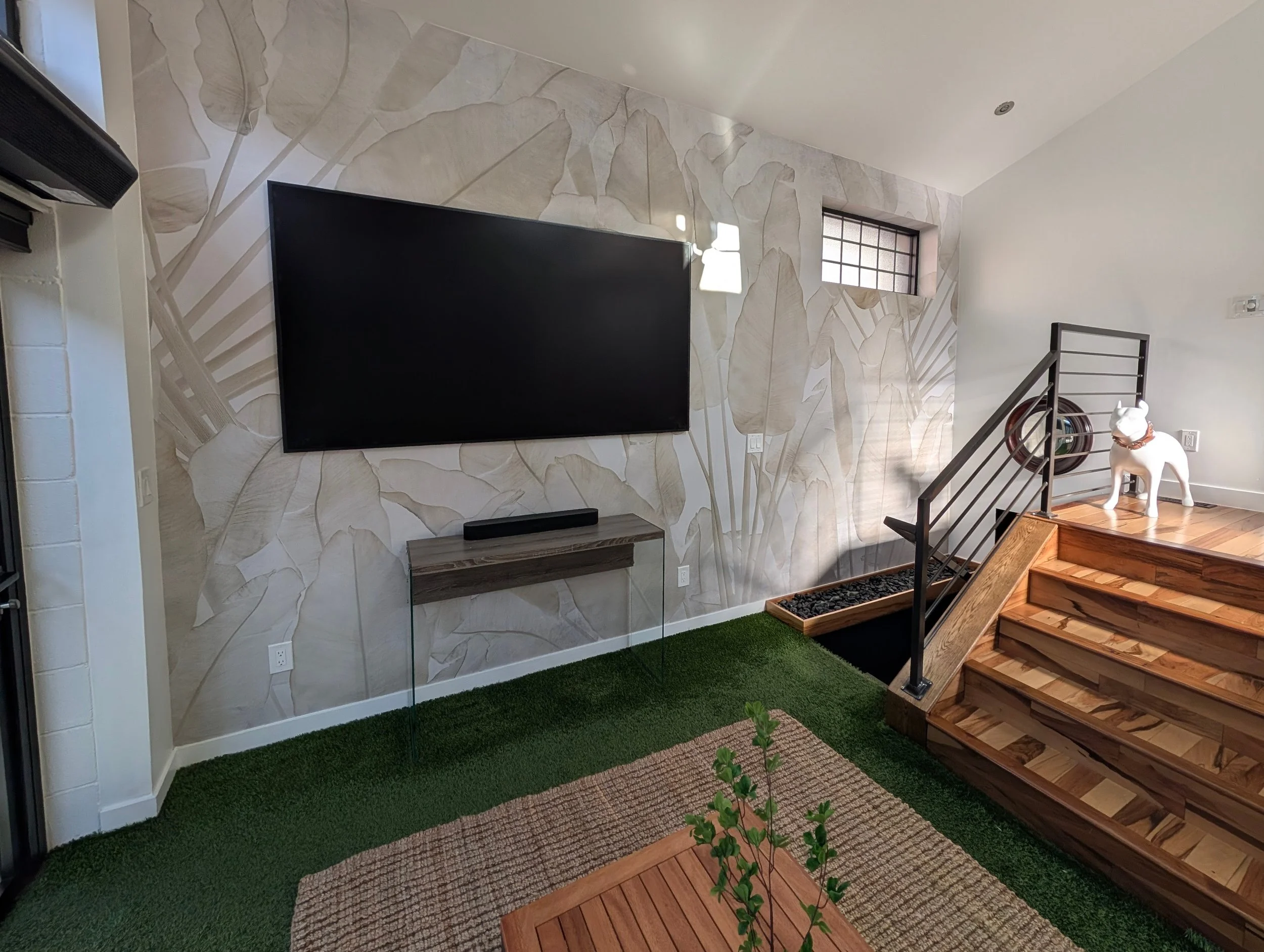 Living room wall with large floral wallpaper, mounted flat-screen TV, small floating wooden shelf below the TV, green carpet, wooden stairs with black metal railing, and a ceramic dog statue on the staircase landing. Small frosted window and a wall-mounted light fixture are also visible.