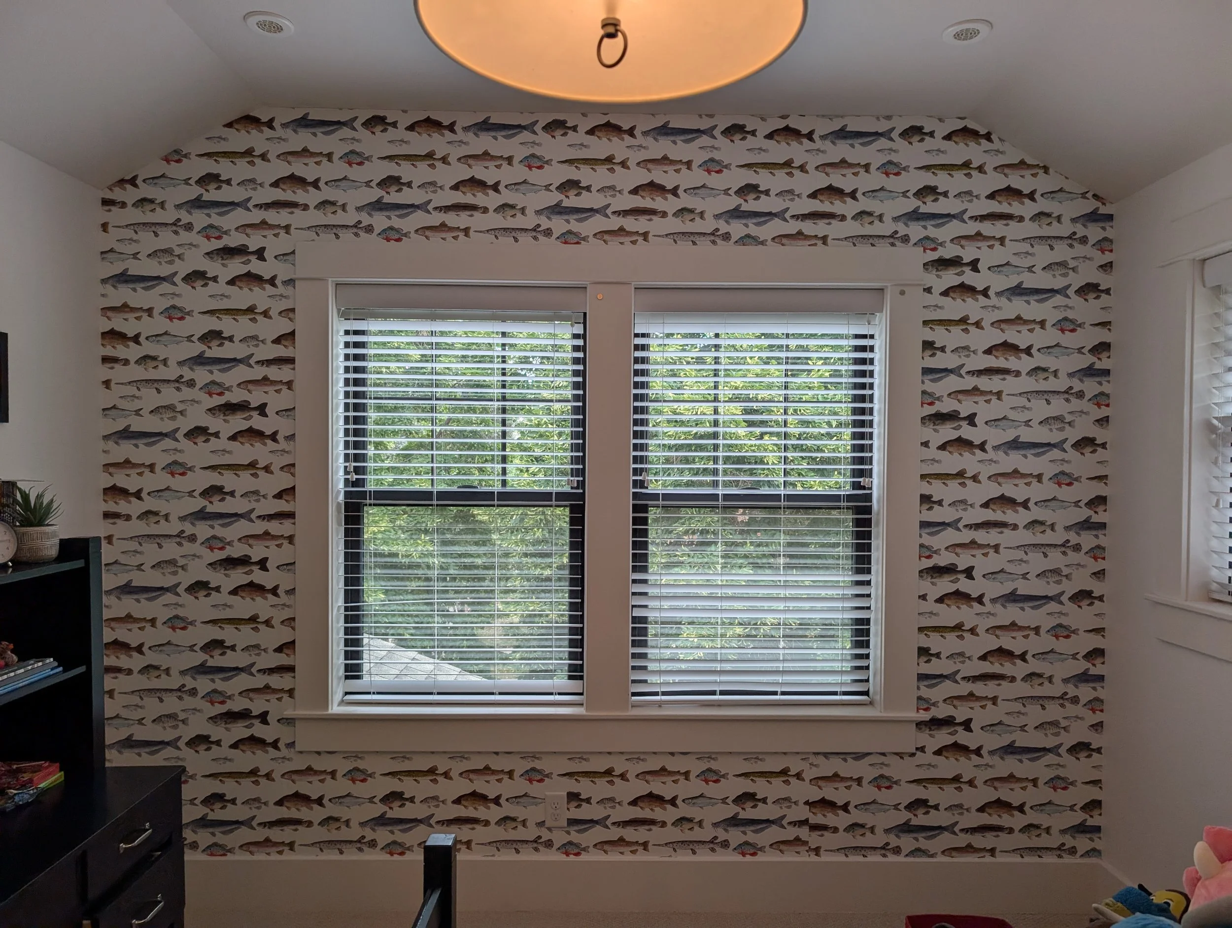 A room with fish-themed wallpaper, white window blinds, and two large windows showing greenery outside.