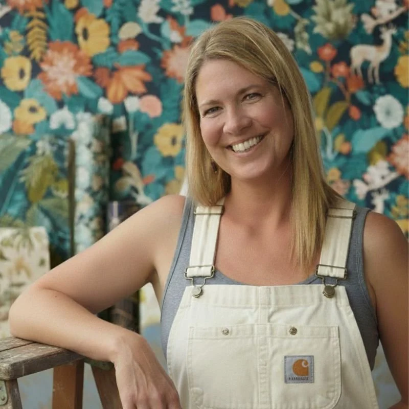 A smiling woman with blonde hair, wearing a gray shirt and white overalls, leaning on a wooden surface in front of a colorful floral patterned background.