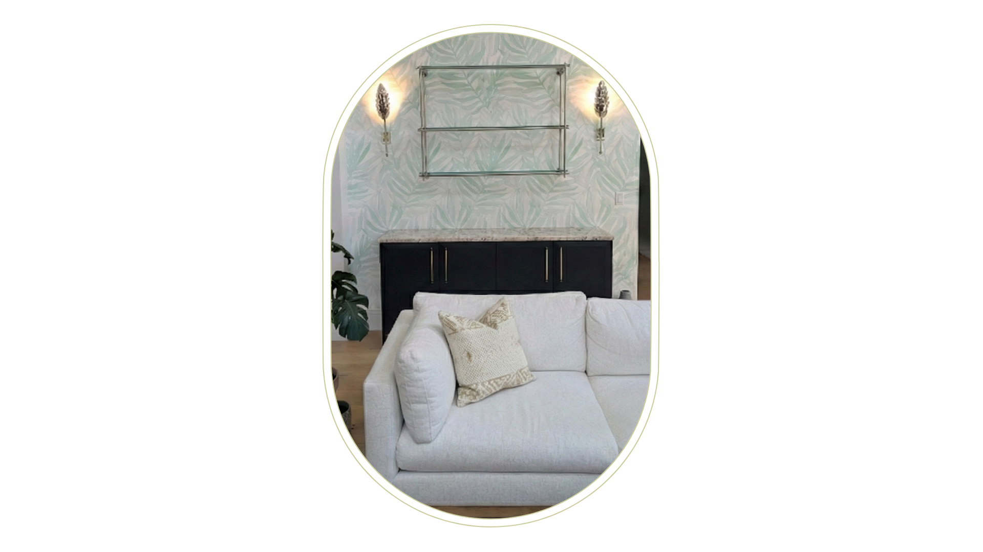 Living room with white sofa and a decorative pillow, black sideboard, peel & stick wallpaper with leaf pattern, two wall-mounted lights, and a metal shelving unit on the wall.