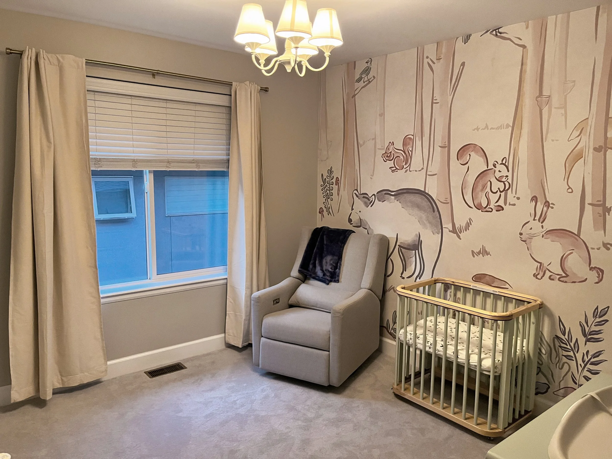 A cozy nursery with a beige armchair, a wooden baby crib, a wall mural featuring forest animals like bears, rabbits, squirrels, and birds, a chandelier with multiple lamps, and a window with cream curtains and a blind.