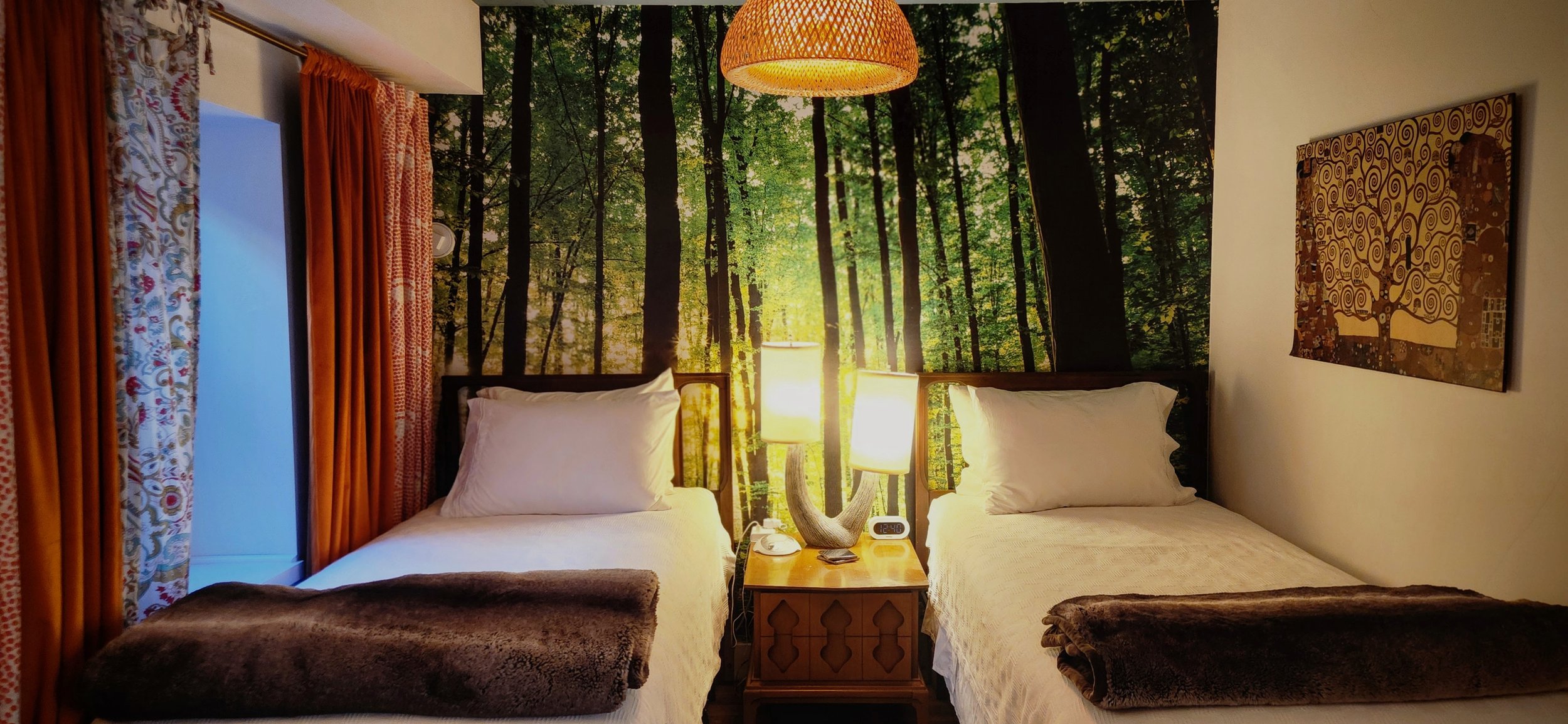 Hotel room with two beds, forest-themed mural on the wall, bedside table with lamps and alarm clock, curtains, and artwork.