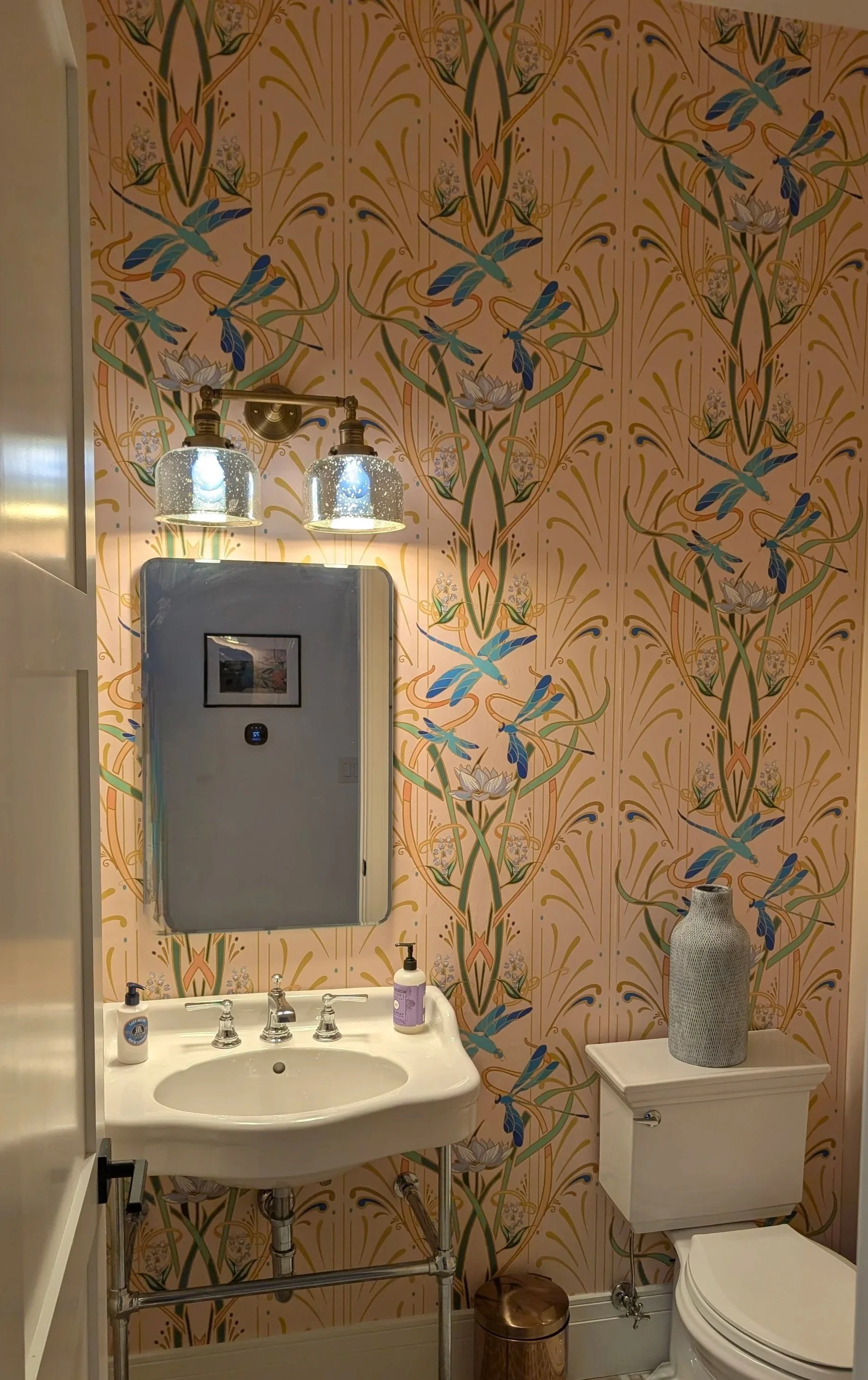 A bathroom with pink floral and bird wallpaper, featuring a white ceramic sink with silver faucets, a wall-mounted mirror, a soap dispenser, wall-mounted light fixtures with glass shades, a white toilet with a gray vase on top, a small trash can, and a partial view of a shower enclosure.