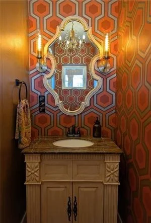 Bathroom with geometric patterned orange wallpaper, a vintage-style mirror, wall sconces, a granite countertop with a sink, black faucet, and a soap dispenser.