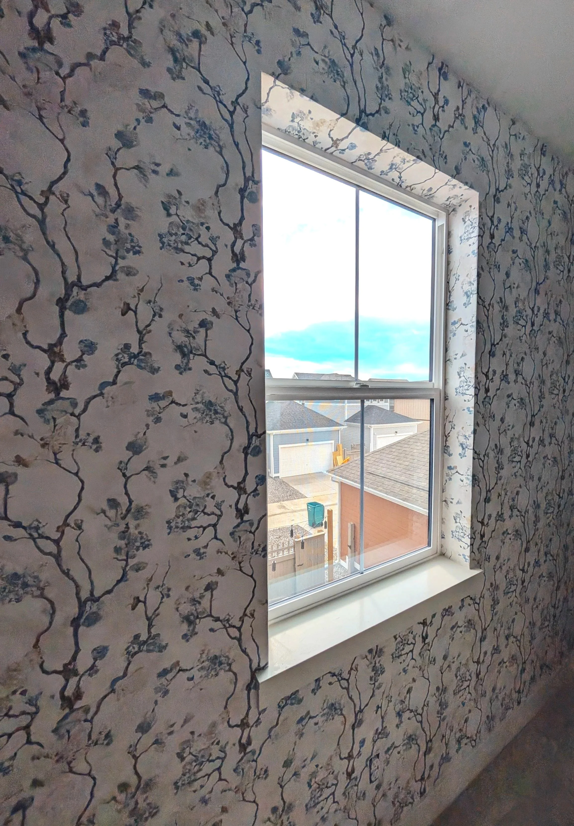 A room with floral wallpaper around a window showing houses and a cloudy sky outside.
