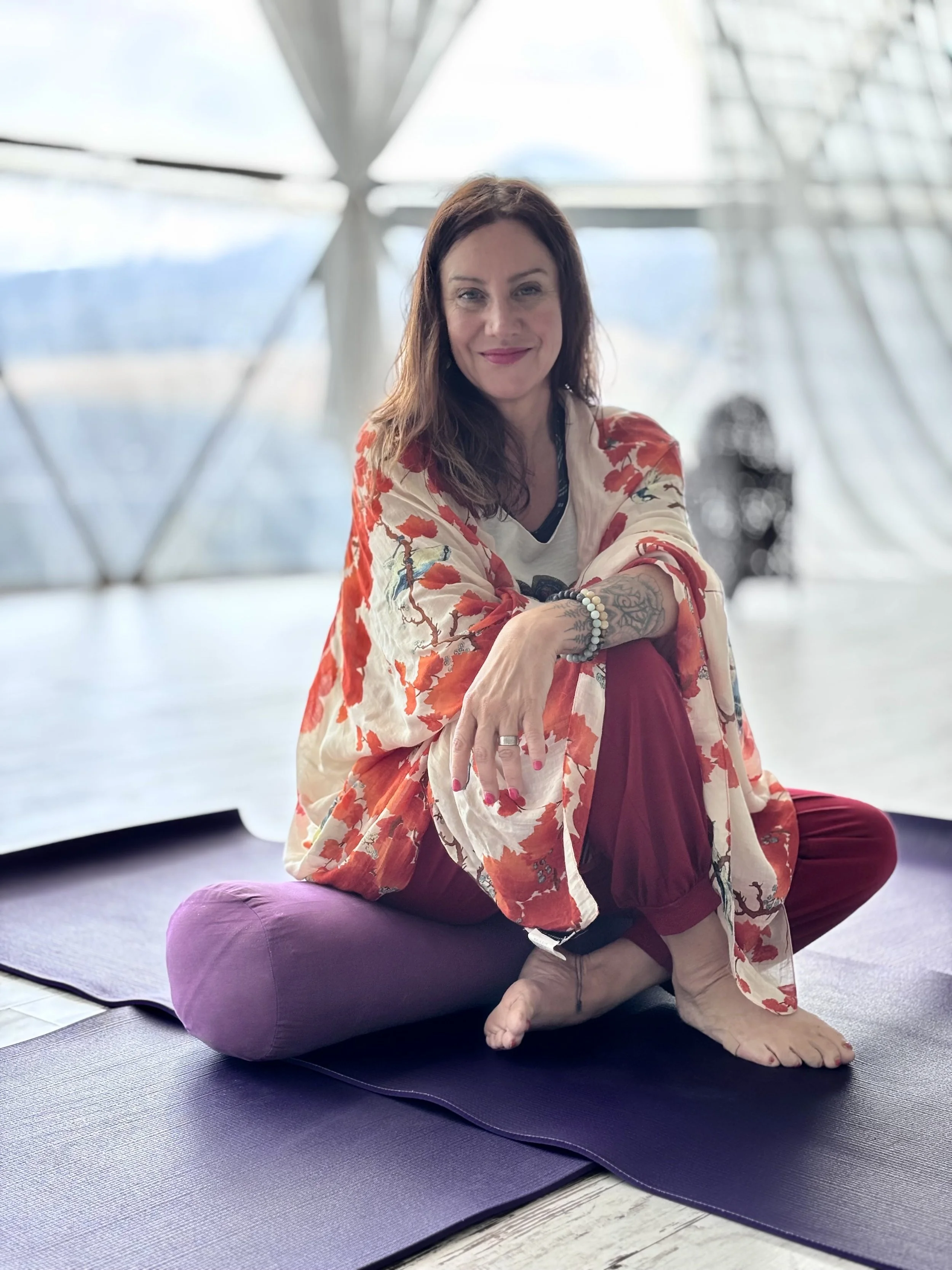 Woman practicing yoga indoors, sitting cross-legged on a yoga mat with windows and a modern building structure in the background.
