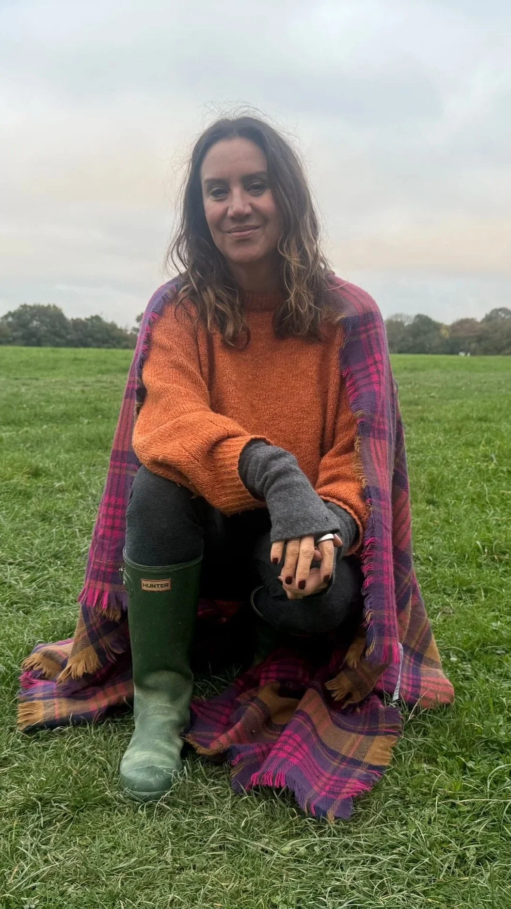 A woman sitting on a grassy field, wearing a multicolored sweater, green rain boots, and a plaid blanket draped over her shoulders. She has curly brown hair and is smiling softly.