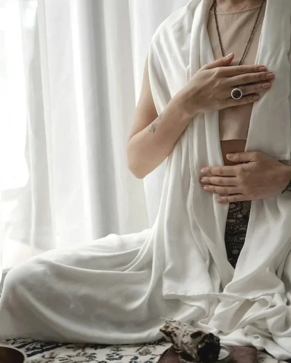Person sitting on a patterned rug in front of white curtains, partially covered in white fabric, holding one hand over their chest, with visible jewelry and tattoos.