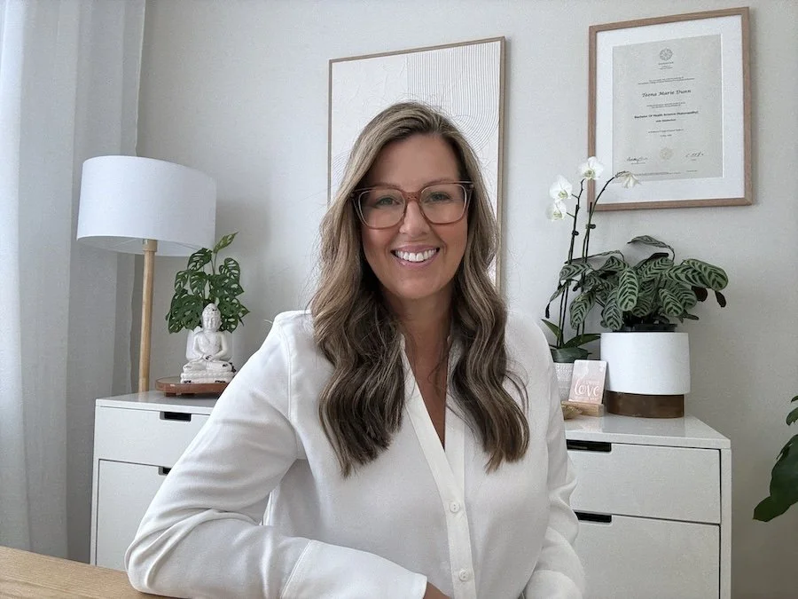 A woman with glasses and long, wavy brown hair smiling at the camera in an office or home workspace. She is wearing a white blouse. Behind her are framed documents, a white cabinet with potted plants, and a lamp.