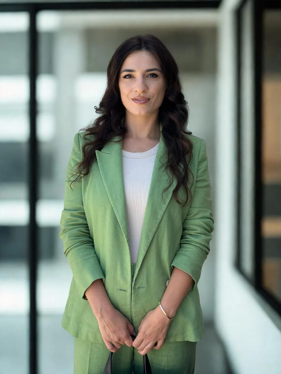 Mujer morena con americana verde claro y top blanco, de pie en un interior con cristales y arquitectura moderna de fondo.