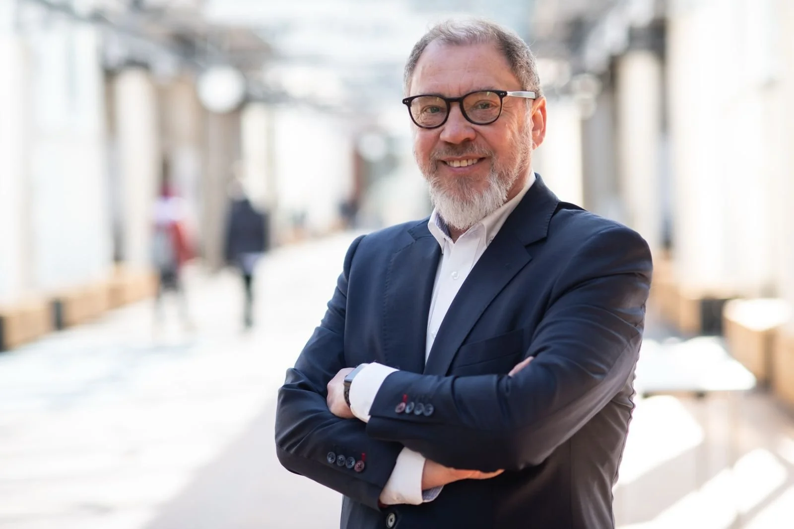 Un hombre con gafas y barba se toma un selfie junto a un río con edificios históricos y una aguja de iglesia de fondo en un día parcialmente nublado.