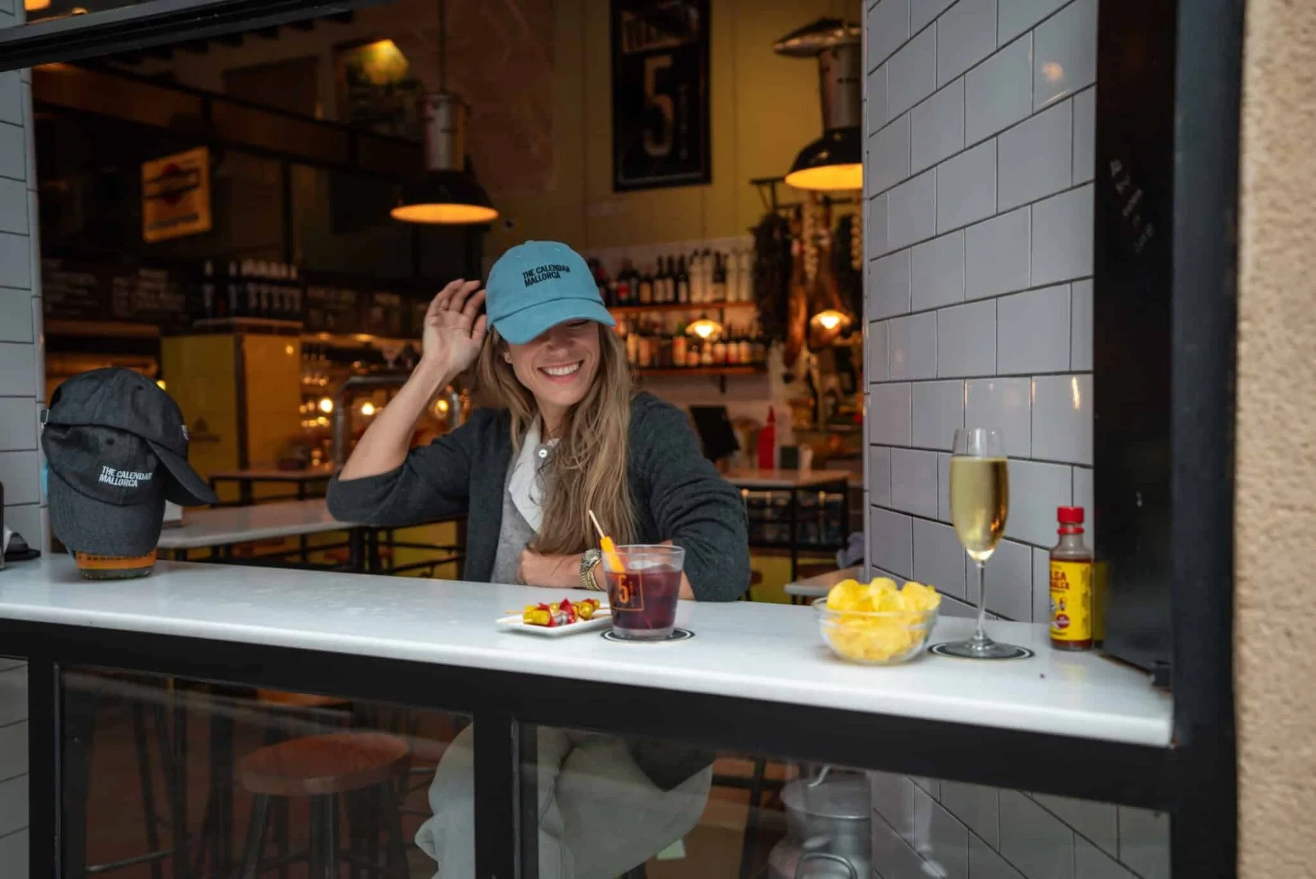 A woman with wavy dark hair wearing a light green blazer and white shirt, standing inside a modern building with glass walls.