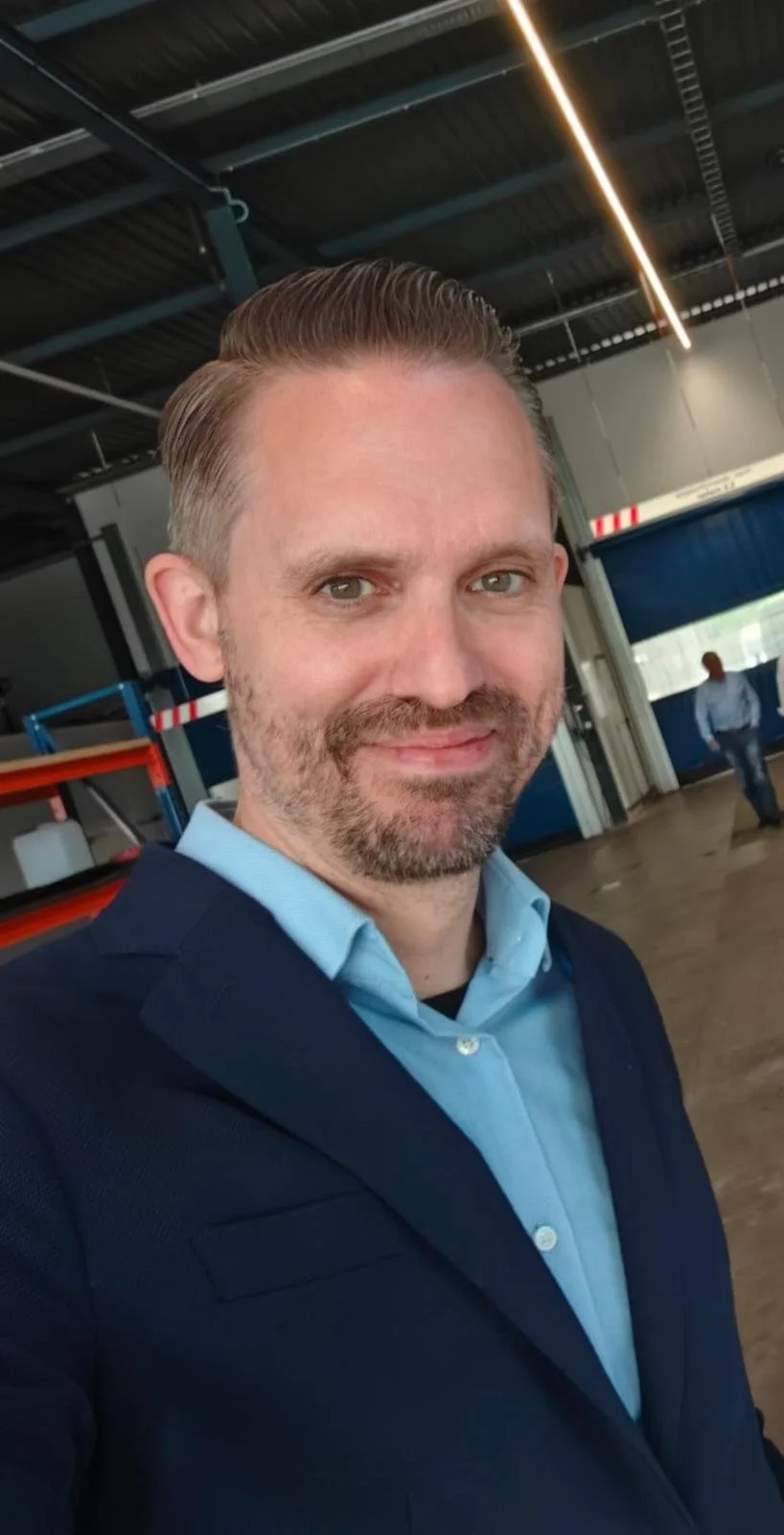 Un hombre con el pelo peinado y barba tomándose un selfie, vestido con un traje de chaqueta oscuro y una camisa azul claro, en un entorno industrial o tipo almacén con techos altos, vigas metálicas y otras personas al fondo.