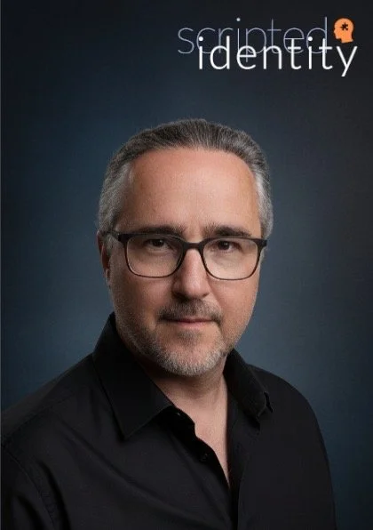 Retrato de un hombre con gafas y pelo canoso delante de un fondo oscuro con las palabras "scripted identity" en la parte superior.