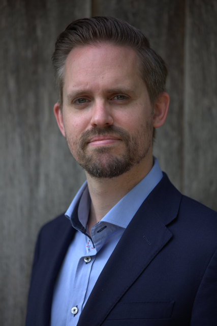 A man with styled hair and a beard taking a selfie, wearing a dark suit jacket and a light blue shirt, in an industrial or warehouse-like setting with high ceilings, metal beams, and other people in the background.