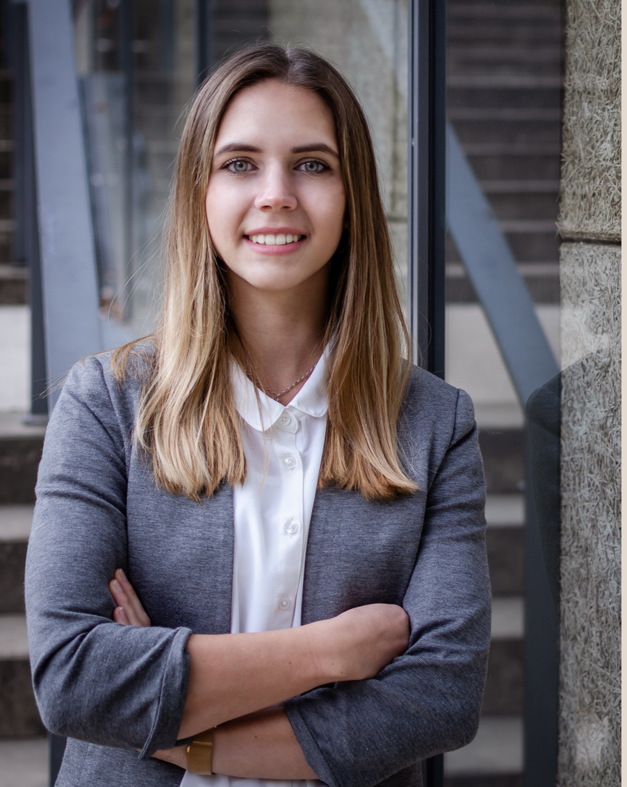 Una mujer joven de pelo rubio y ojos azules sonríe, lleva una americana gris sobre una camisa blanca y está de pie en el exterior, cerca de una ventana de cristal y un muro de piedra.