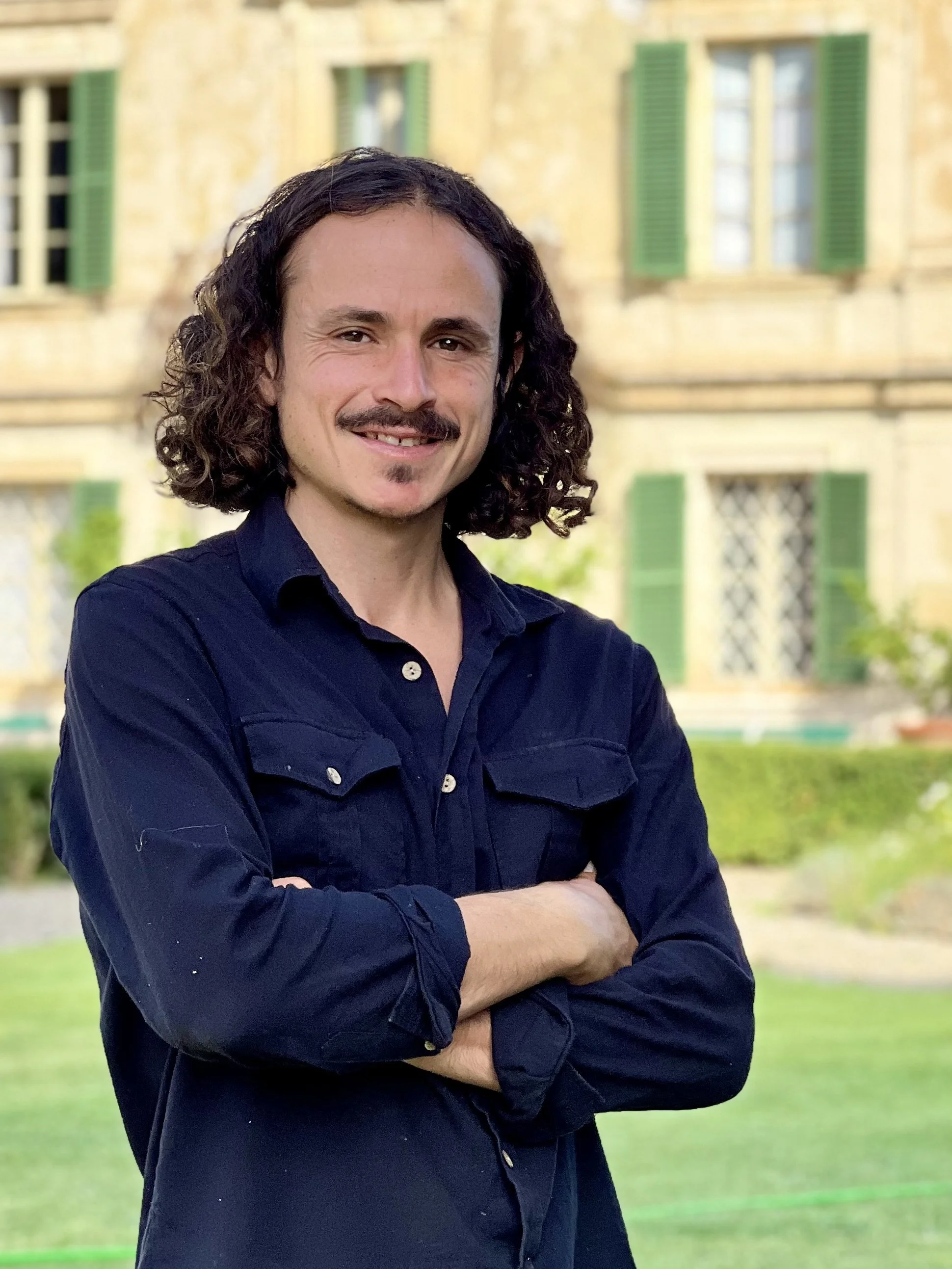 A man with curly dark hair, a mustache, and a goatee, smiling with arms crossed, standing outdoors in front of a building with green shutters and a garden.