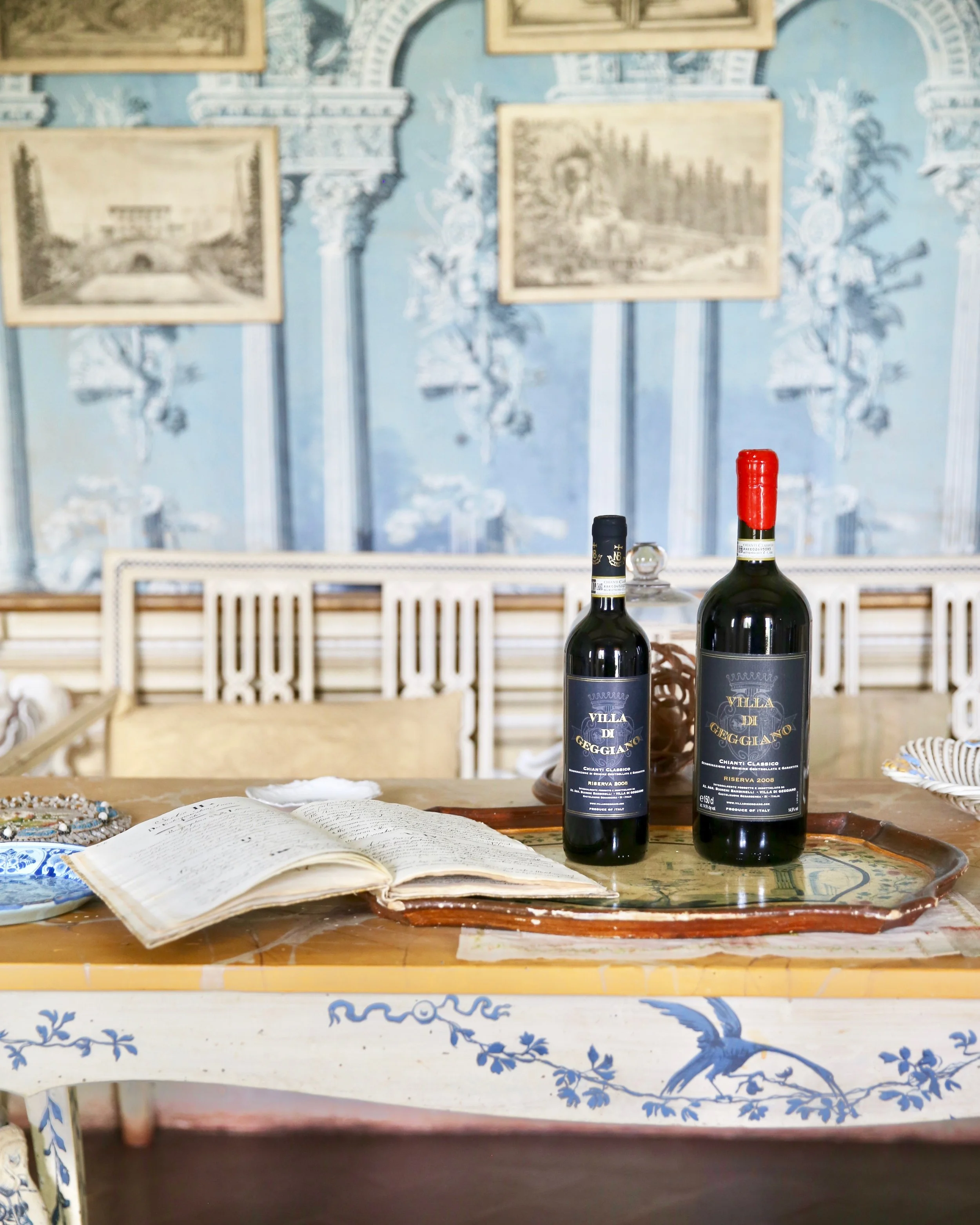 A table with two bottles of Villa di Geggiano Chianti wine, an open book, and a decorative tray. The background features a blue and white wall with framed artwork.
