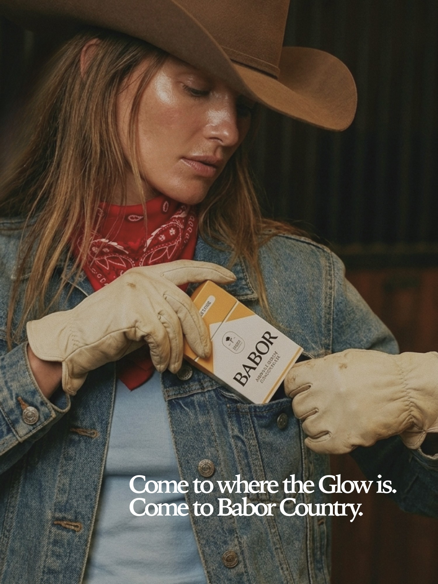 A woman wearing a cowboy hat, a red bandana, a denim jacket, and gloves, holding a box labeled 'BABOR' in a retail or promotional setting.