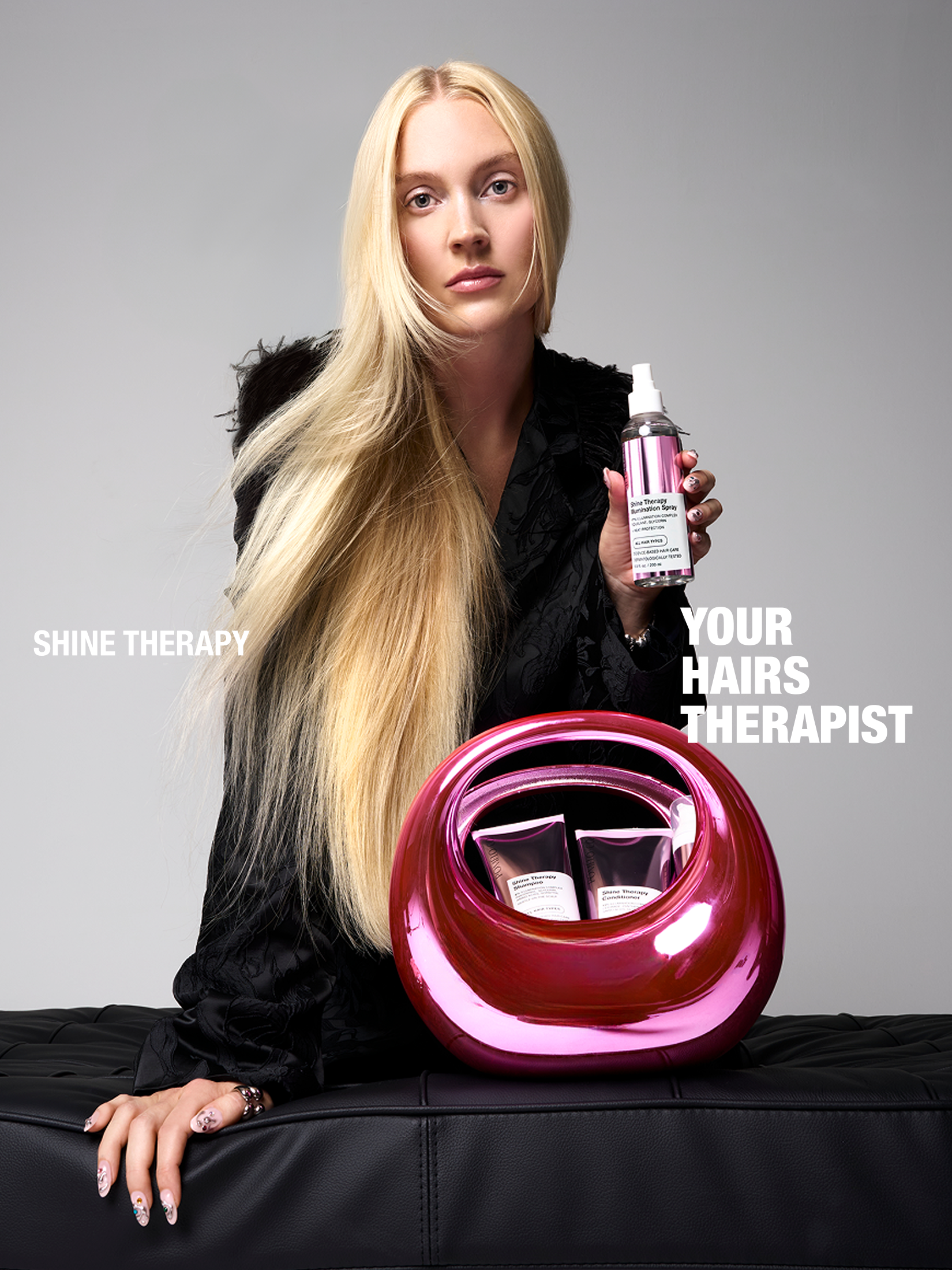A blonde woman with long hair holding a spray bottle of Shine Therapy while sitting on a black leather surface, with a pink reflective object in front of her and text overlaid on the image that reads 'SHINE THERAPY' and 'YOUR HAIR'S THERAPIST'.
