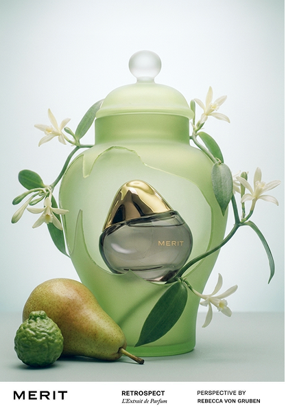 A perfume bottle labeled 'MERIT' with a green and gold design, surrounded by white flowers, a pear, and a small green gourd on a white background.