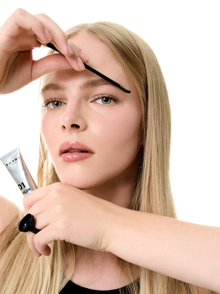 A woman applying eyebrow makeup with a small brush and holding a tube of gel.