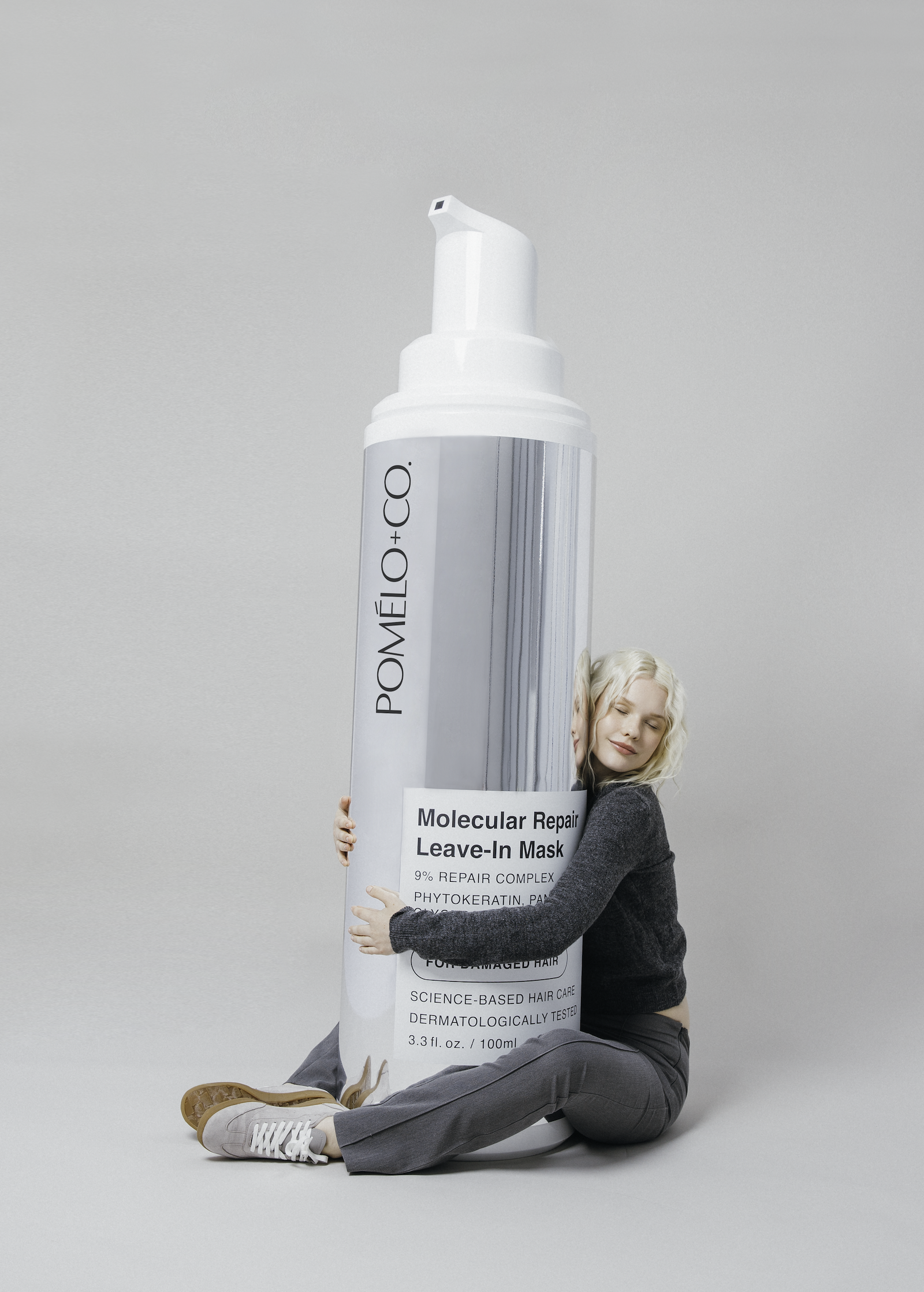 A woman sitting on the floor hugging a large tube of molecular repair leave-in mask, with her eyes closed and a content expression.