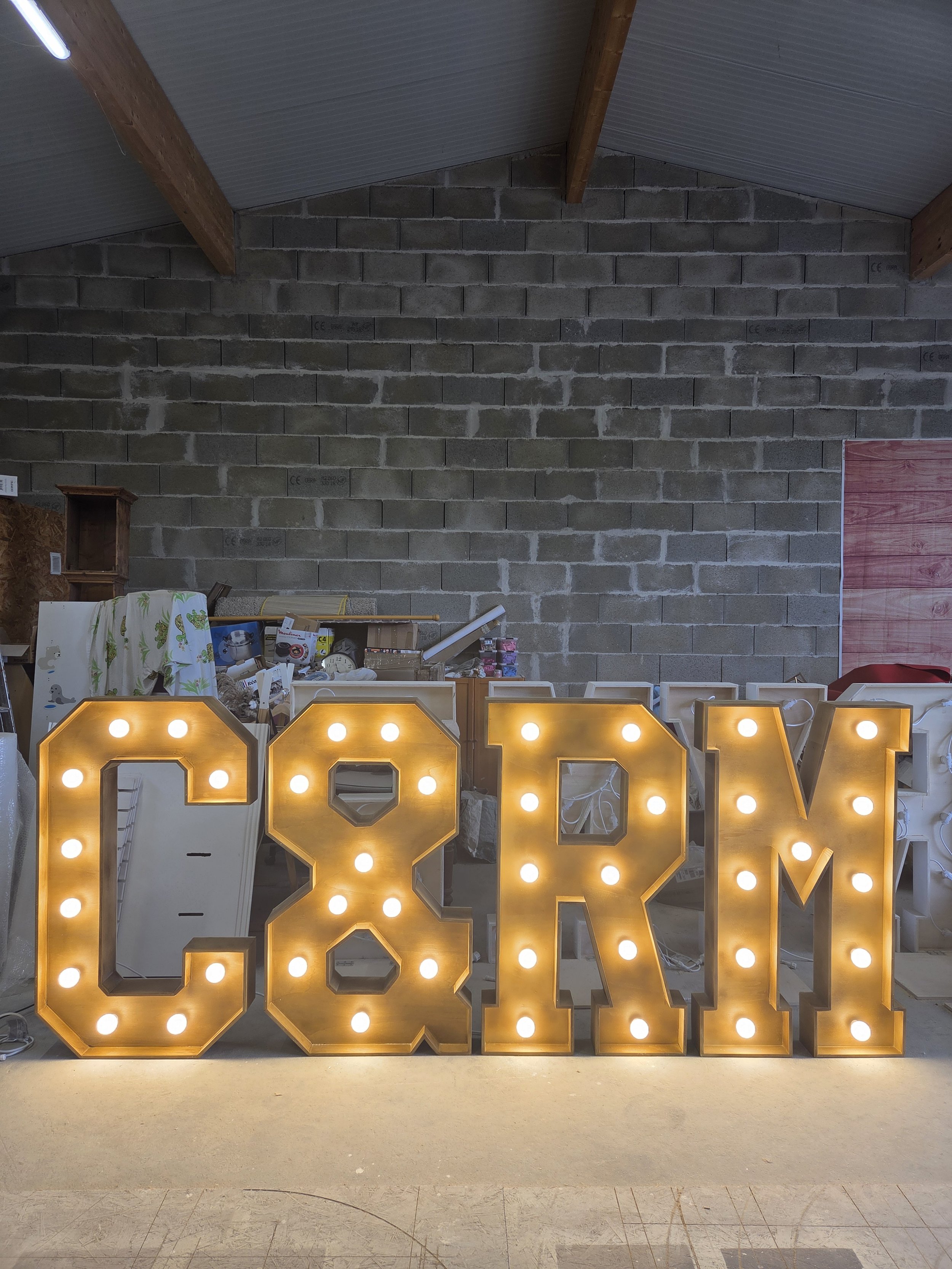 Lettres lumineuses formant 'C & R M' sur un sol, dans un espace de stockage ou atelier, avec un mur en briques et un plafond en bois.