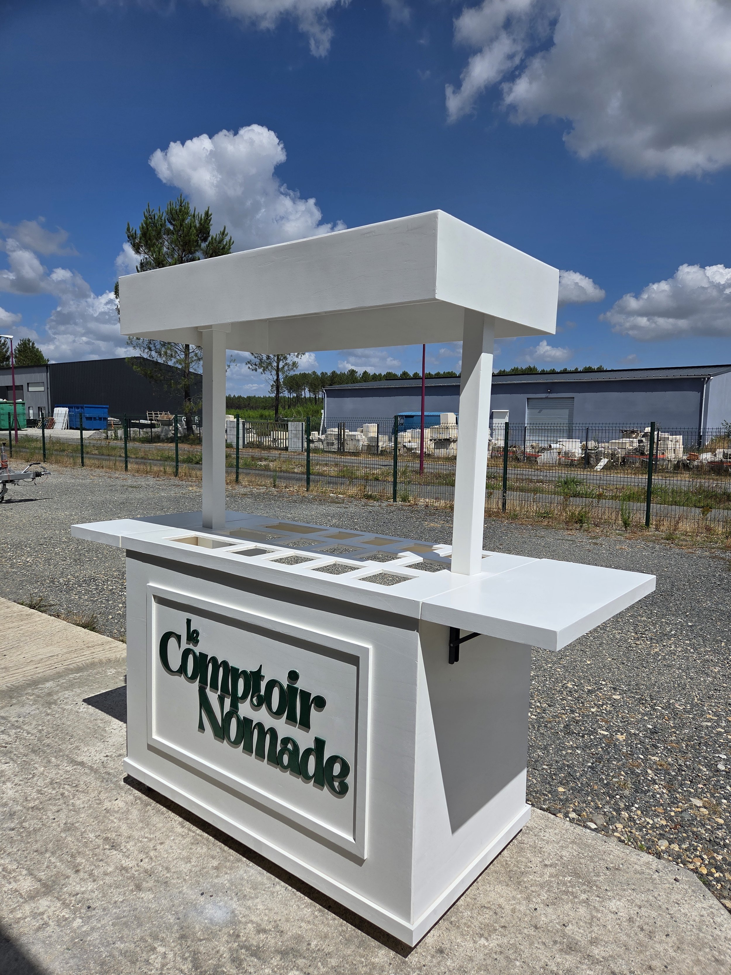 Un stand blanc en forme de kiosque avec un panneau indiquant 'le Comptoir Nomade' placé en dehors d'un entrepôt ou d'une zone industrielle sous un ciel en partie nuageux.