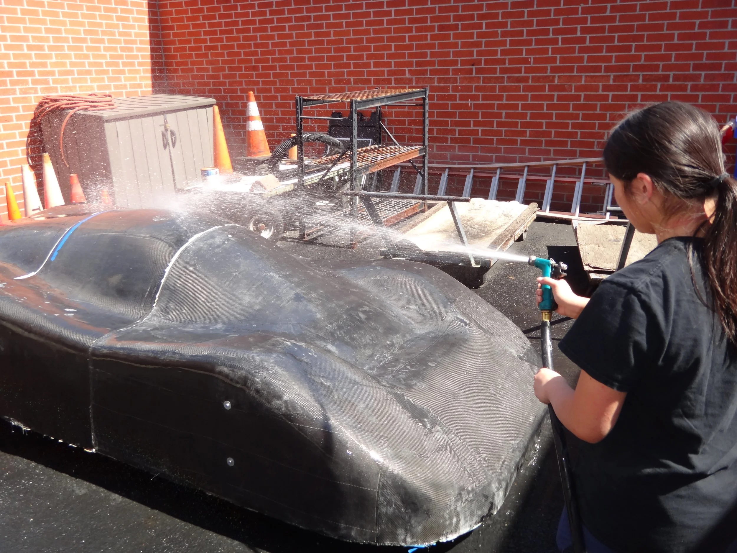 A person with dark hair tied back in a ponytail washing a black race car with a hose, spray of water and foam on the car's surface, in front of a brick wall and orange traffic cones.