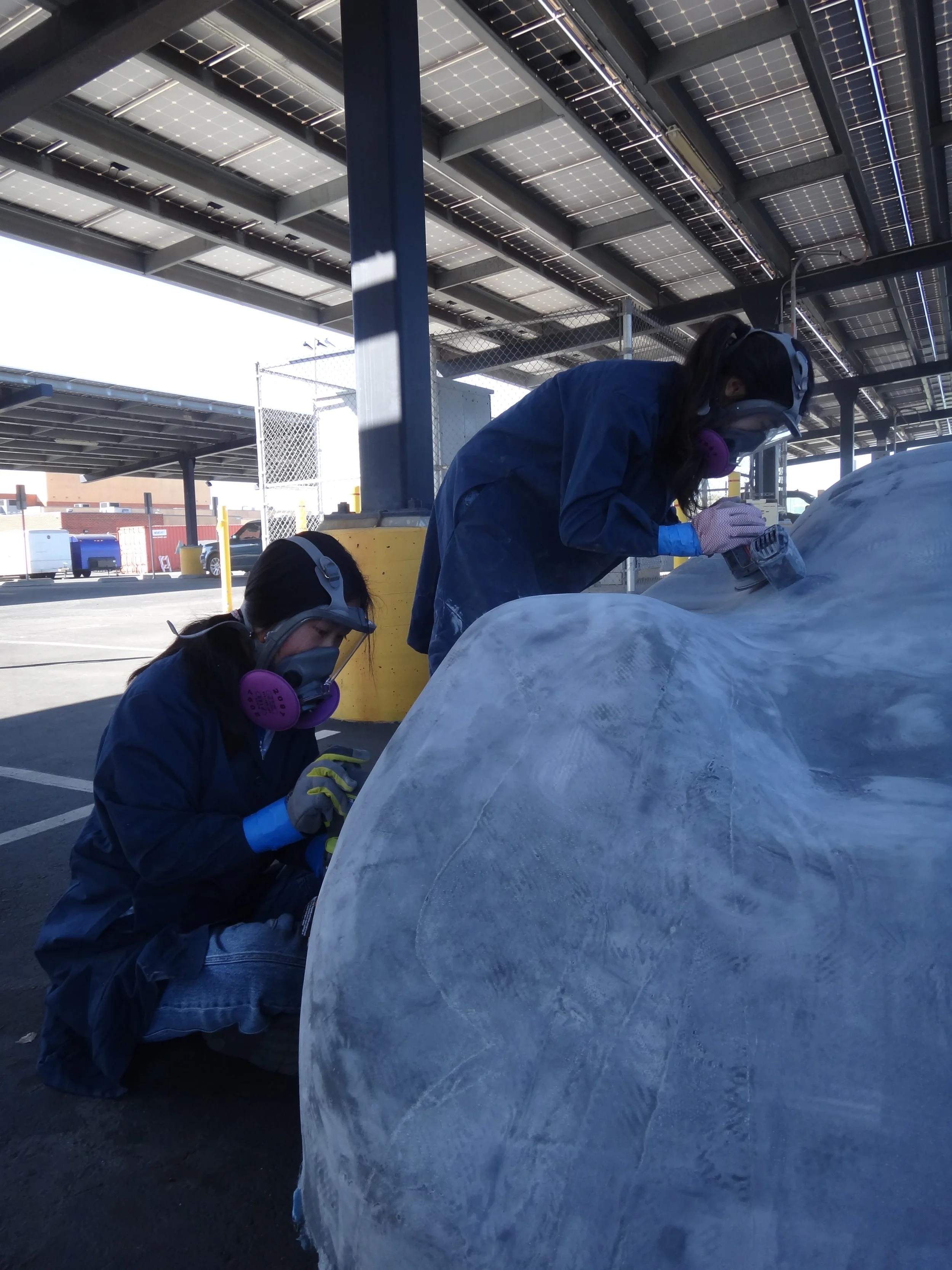 Two individuals wearing protective masks, gloves, and blue coveralls working on a large, rounded block of ice with power tools in an outdoor parking area under a solar panel-covered structure.