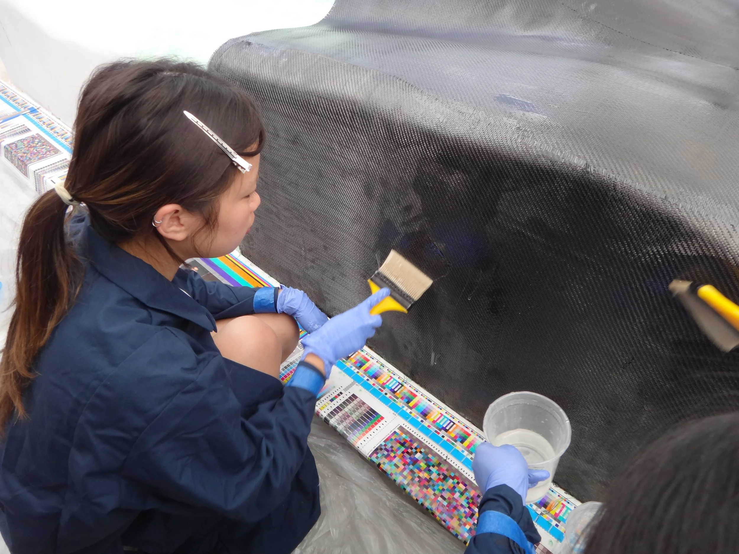 A woman wearing a blue jacket and gloves painting a black carbon fiber surface with a paintbrush, sitting on the floor next to a colorful test pattern sheet and a cup of liquid.