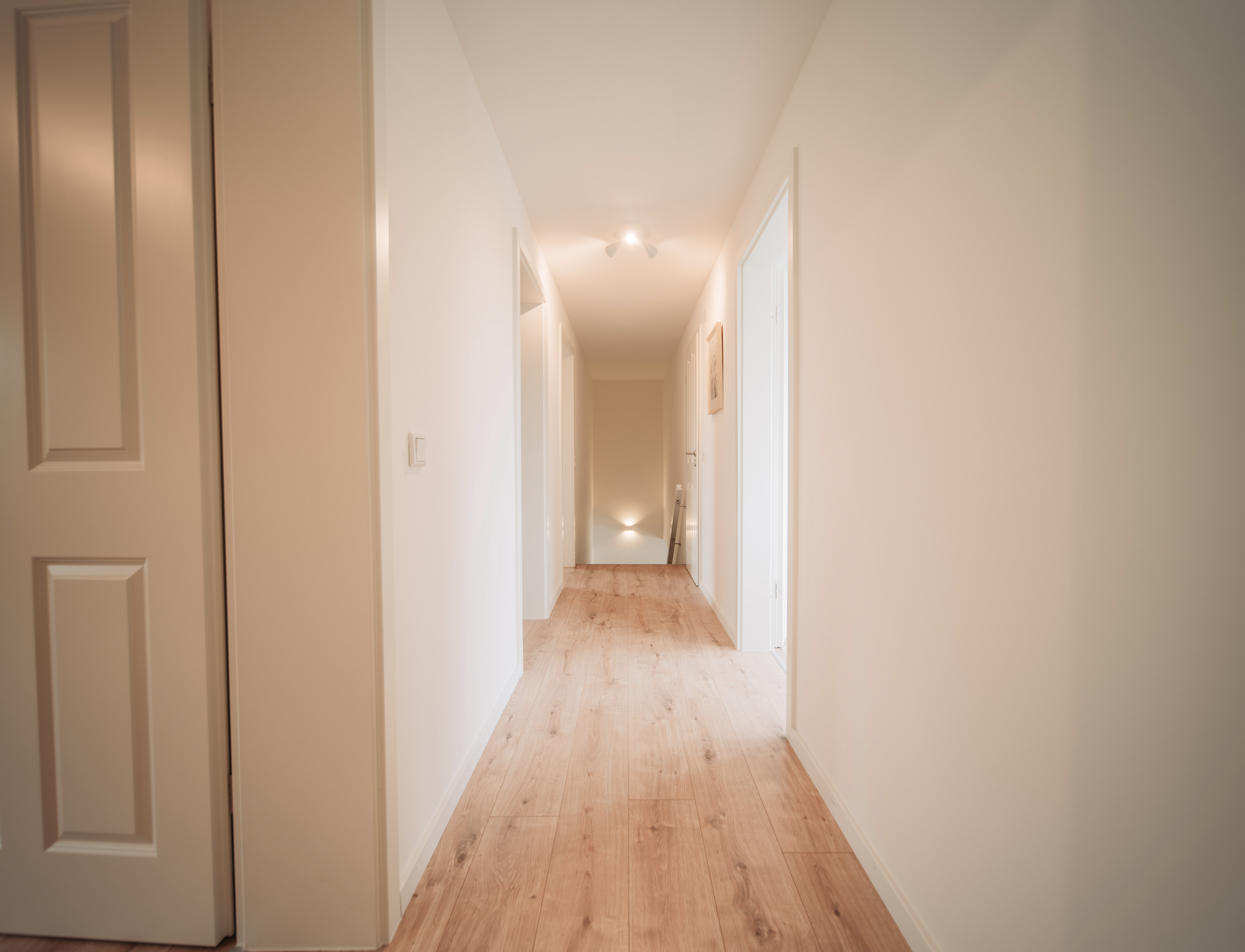 Ein heller Flur in einem Haus mit weißen Wänden, Holzboden und mehreren Türen auf beiden Seiten, und eine Treppe am Ende des Flurs.