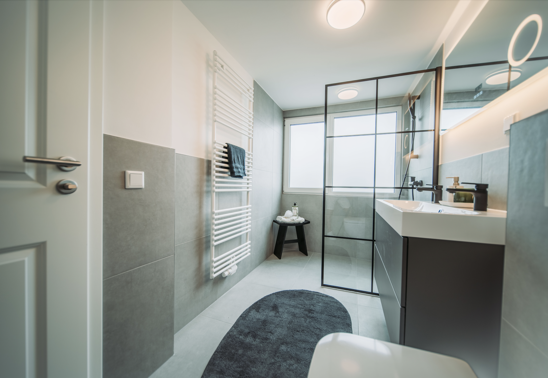 Modernes Badezimmer mit grauen Fliesen, Doppelwaschbecken, Spiegel und separater Duschbereich mit Glaswand, schwarzer Hocker, Handtücher und Hygieneprodukte auf Ablage