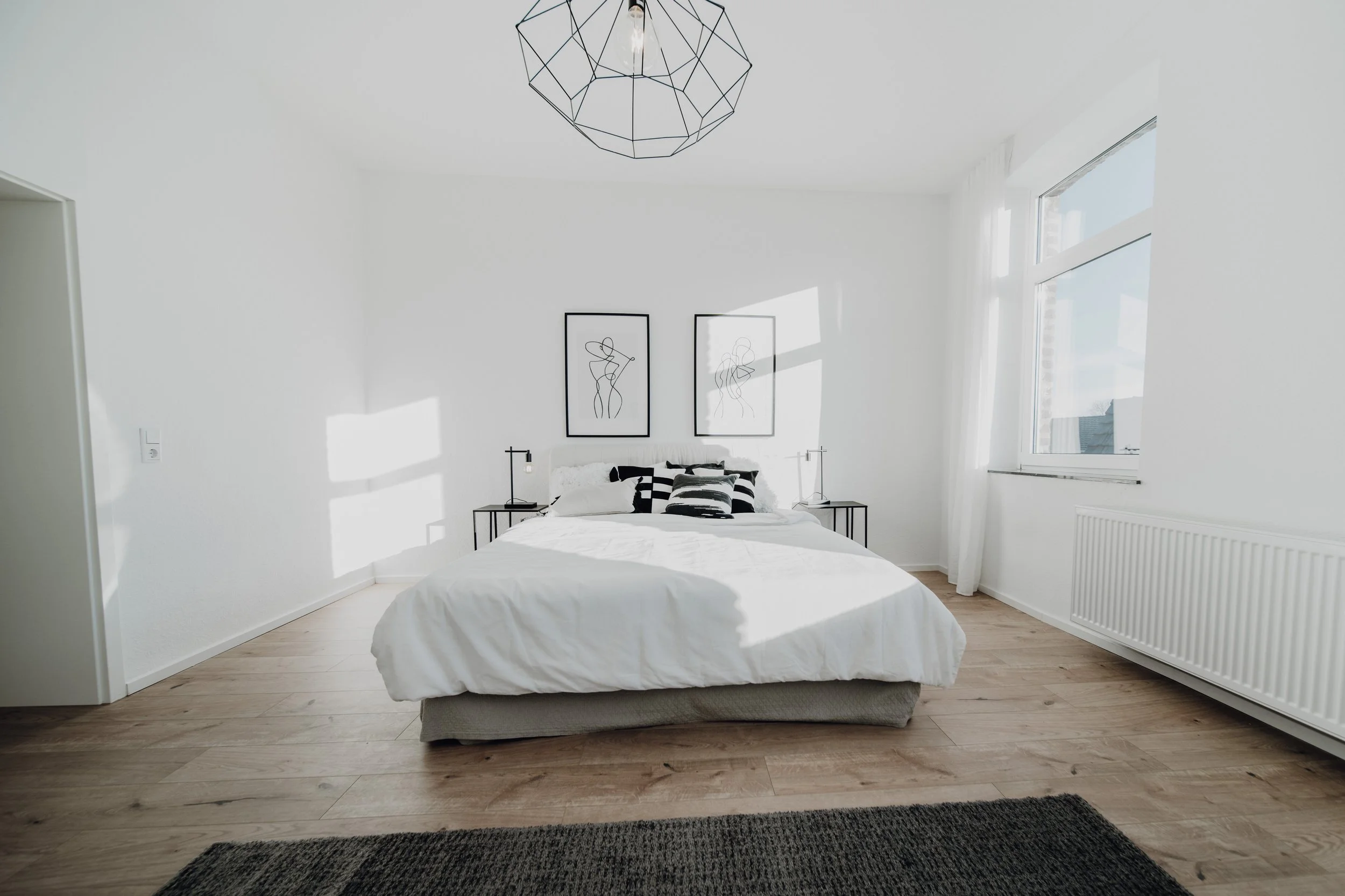 Ein schlichtes, weißes Schlafzimmer mit einem Bett, schwarzen und weißen Kissen, Wandbildern mit Linienzeichnungen, Seitentischen mit Lampen, Holzfußboden, einem großen Fenster mit weißen Vorhängen und einer Deckenleuchte.