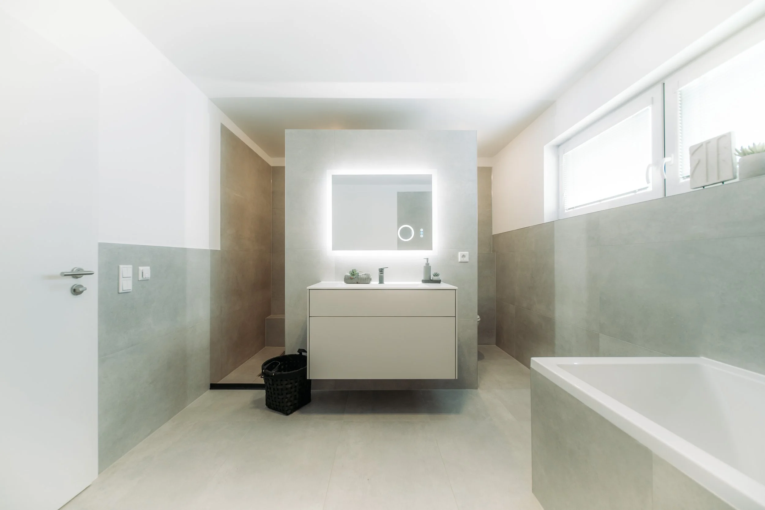 Moderner, minimalistisch gestalteter Badezimmerraum mit Waschbecken, beleuchtetem Spiegel, Badewanne und natürlichem Licht durch ein Fenster.