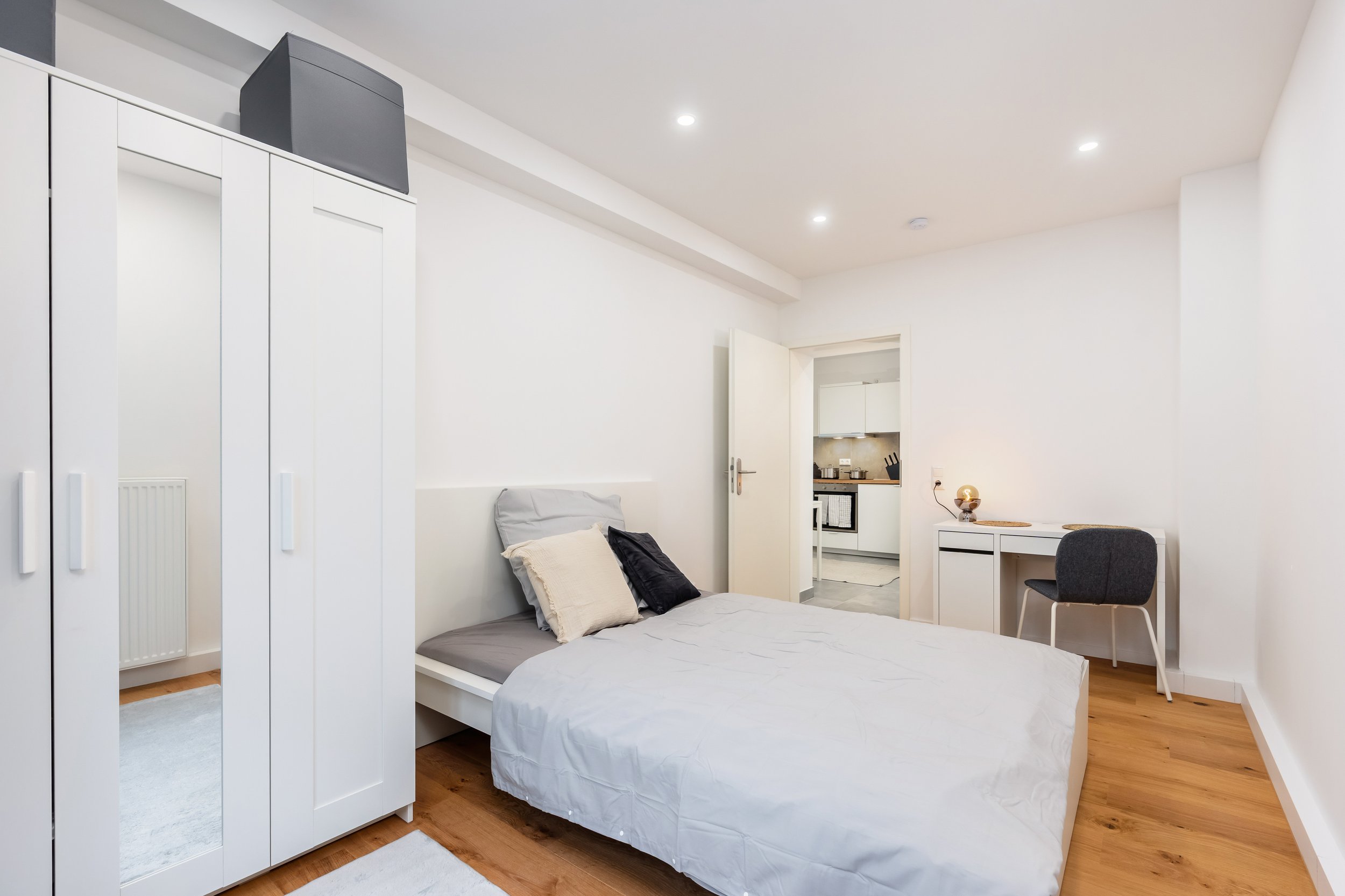 Schlafzimmer mit weißem Bett, grauen und schwarzen Kissen, weißen Wänden, Holzboden, Kleiderschrank, Schreibtisch mit grauem Stuhl, offener Tür zum Küchenbereich.