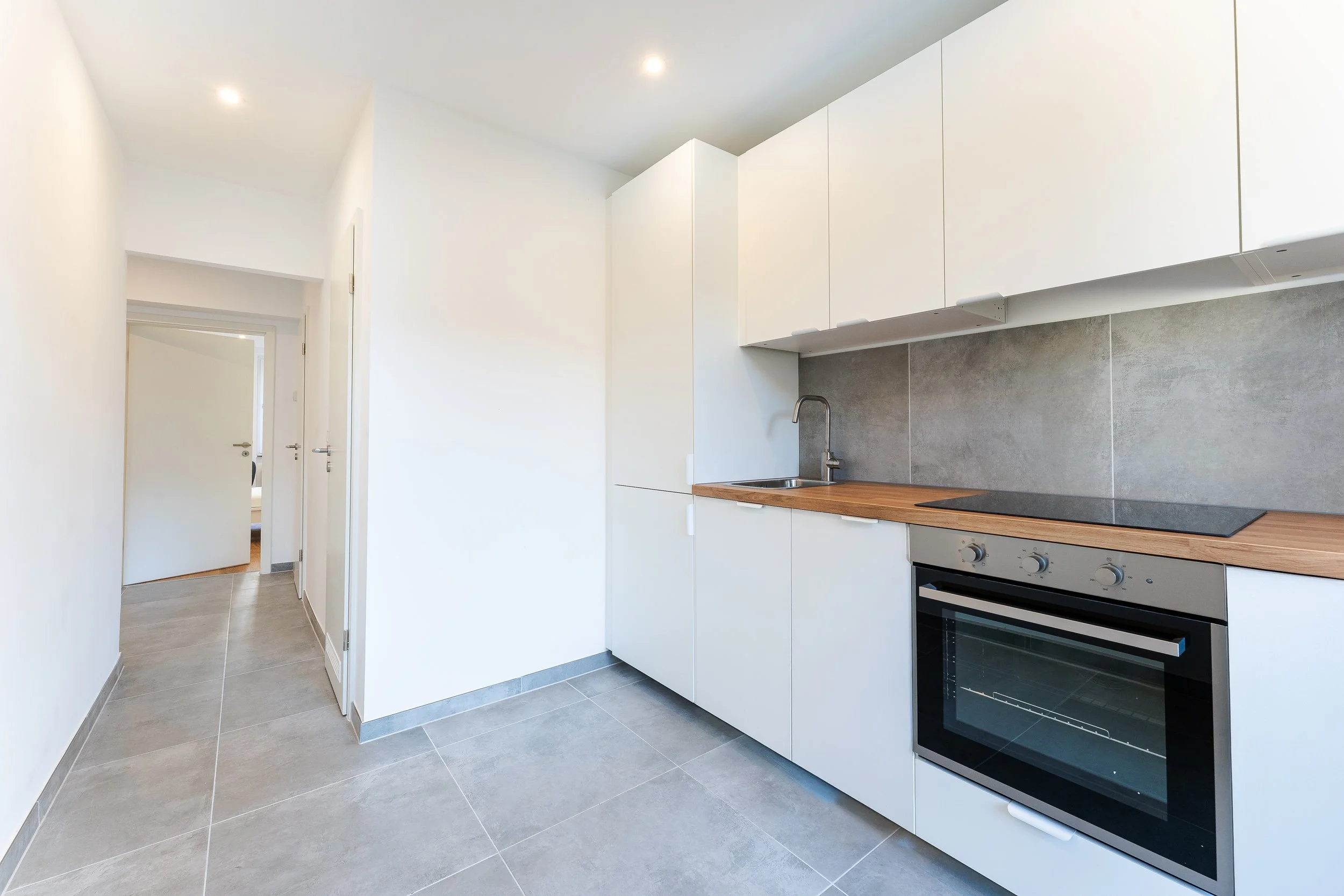 Moderne, kleine Küche mit weißen Schränken, Holzarbeitsplatte und Backofen, grauer Fliesenspiegel und Fliesenboden.